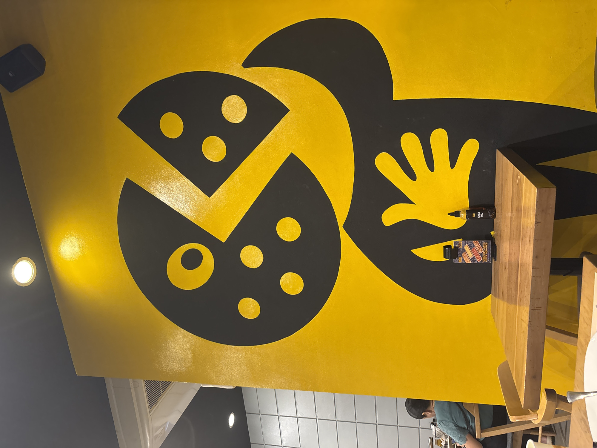 The image depicts a brightly lit interior, likely a restaurant, with a prominent yellow wall featuring a large black and yellow graphic of a stylized figure holding a slice of pizza. A wooden table and chairs are visible in the foreground, with a few bottles of condiments on the table. In the background, a person is seated at another table. The overall ambiance suggests a casual dining establishment. This image was taken at Yellow Cab Pizza in SM Lanang Premier, Davao City, Philippines.