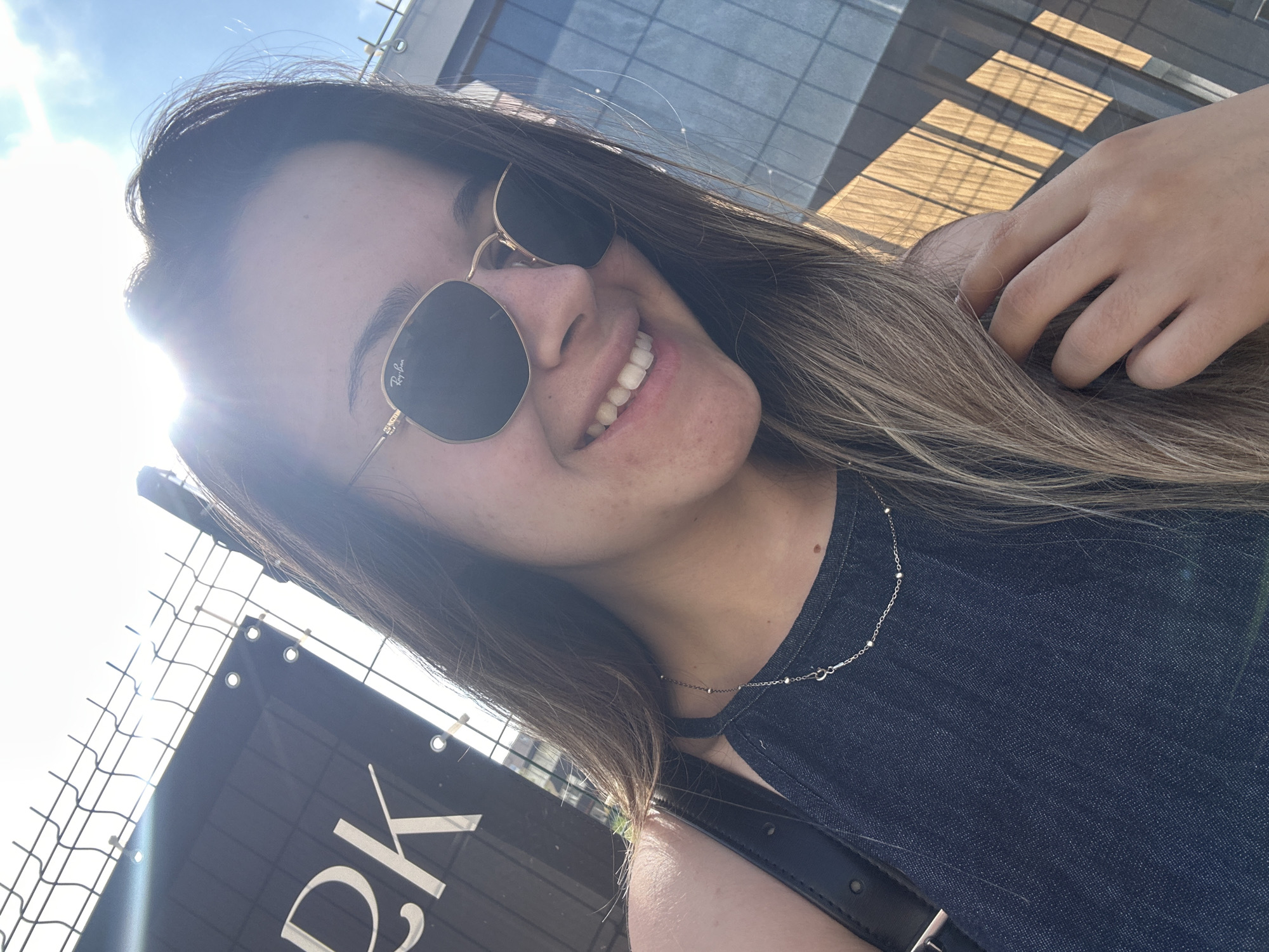 The image captures a bright, sunlit outdoor selfie of a young woman from a slightly low angle. The woman has long, light brown hair and is smiling broadly, revealing her teeth. She wears dark, hexagonal sunglasses with "Ray-Ban" visibly inscribed on the lens. A prominent sun flare emanates from the upper left, casting a strong glow across her face and hair.
She is dressed in a dark blue, sleeveless top, possibly denim, and a delicate silver chain necklace with small beads. Her left hand is casually placed near her head, touching her hair.
The background indicates an urban setting, likely on a sunny day in Sofia, Bulgaria. To the left, a wire mesh fence is present, attached to which is a black banner with white text. The letters "RK" are clearly visible on the banner. Beyond the fence is a bright, clear sky. In the upper right, a modern building with large glass windows and some wooden paneling is visible, reflecting the sunlight. The overall mood conveyed is cheerful and relaxed under bright sunshine.