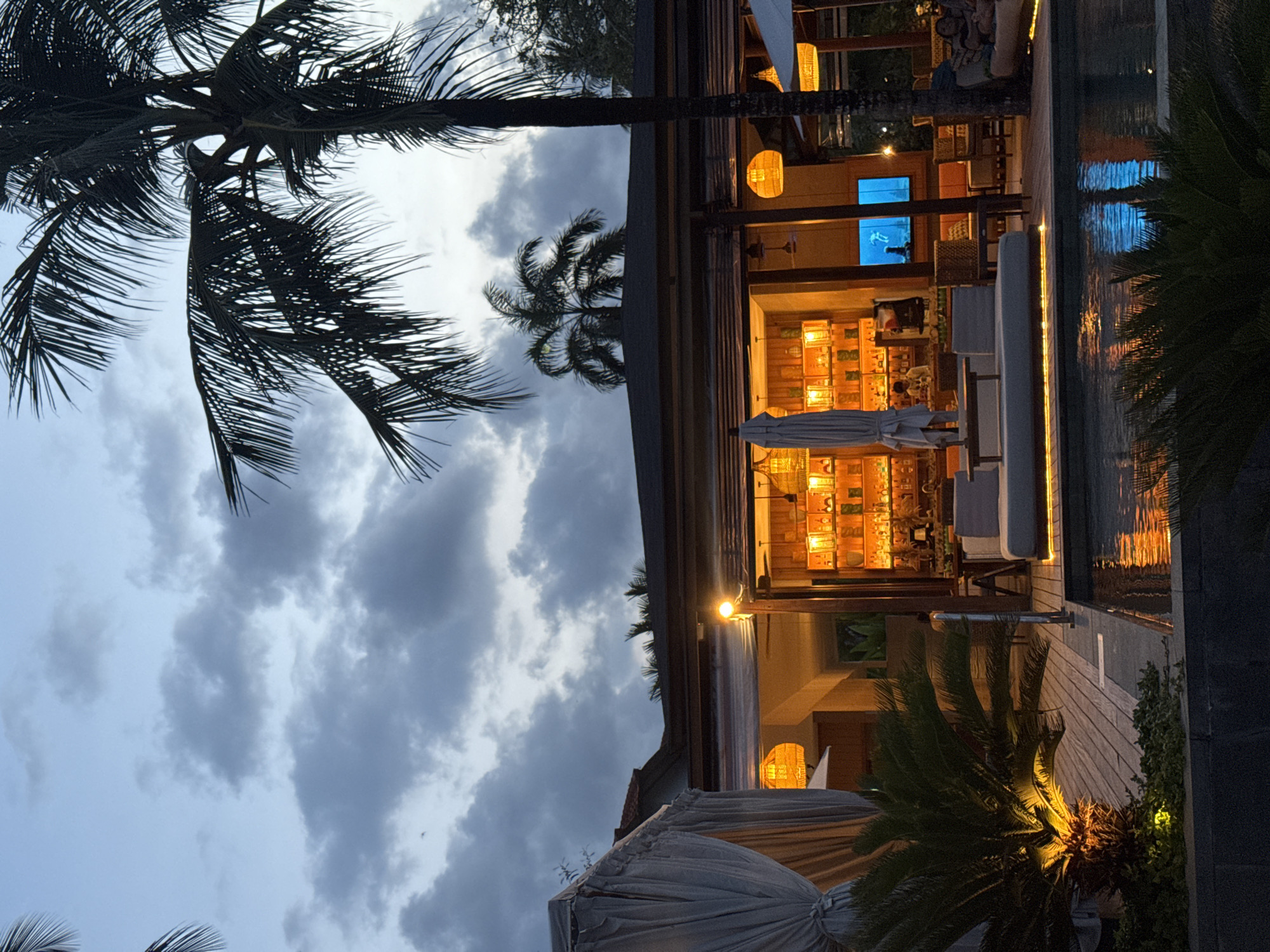 The image, taken from a low angle, shows a partially outdoor bar or restaurant at night in Kuta Selatan, Indonesia. Silhouetted palm trees frame the left side of the frame, juxtaposed against a cloudy sky. To the right, a building with warm internal lighting is visible. A pool or water feature runs alongside the building, reflecting the lights. There are tables, chairs, and white lounge seating visible within the bar area. Hanging wicker light fixtures add to the ambient lighting. A large flat-screen television displays a vibrant blue image. The scene appears still and unoccupied, and the photo's focus is on the architecture and light against the natural elements.