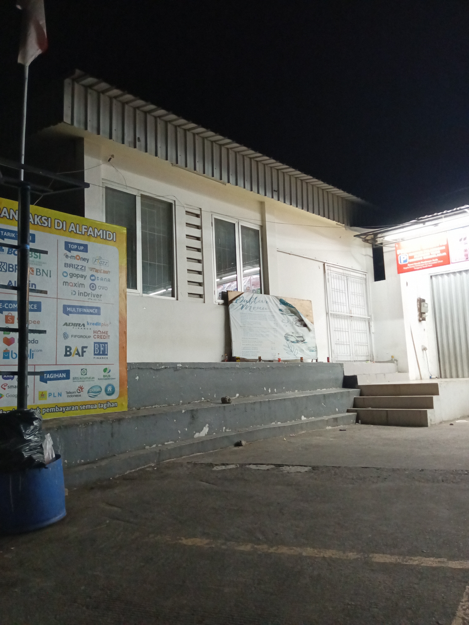 An exterior night-time, eye-level shot depicts the front of a commercial building in Makassar, Indonesia, likely a retail store such as Alfamidi, as indicated by the yellow banner on the left. The building features a white facade with several windows and a corrugated metal roof overhang. A ramp and several concrete steps lead up to the entrance. To the left, a blue trash bin is overflowing with refuse, and a flag pole with a partially visible flag stands nearby. A yellow banner is displayed prominently on the left side of the image, listing various payment and financial services, including "TOP UP e-money", "BRIZZI", "gopay", "DANA", "maxim", "ovo", "inDriver", "ADIRA FINANCE", "kreditplus", "FIFGROUP", "HOME CREDIT", "BAF FINANCE", "BFI FINANCE", "PLN", "Telkom Indonesia", and "bpom". A large, white sign with text and graphics, possibly a menu or advertisement, is propped against the building to the right of the ramp. A red sign with a white "P" indicating parking is visible above a doorway on the right side of the building. The ground in front of the building is paved and appears to be a parking area or walkway.