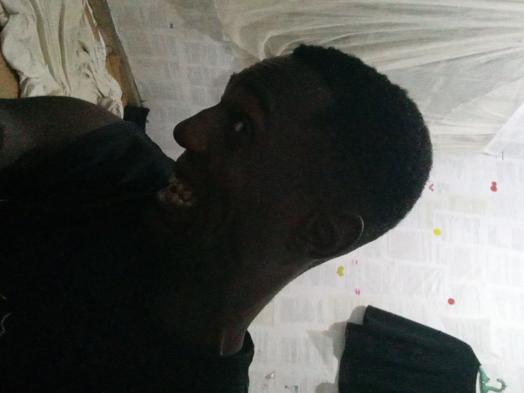 A close-up, angled profile of a dark-skinned man with short black hair is the primary subject. He is wearing a dark shirt and has his mouth open. The background is a white wall with what appears to be a partially visible white cloth or netting. The location cannot be determined to be Akwete, Nigeria, based solely on the provided image.