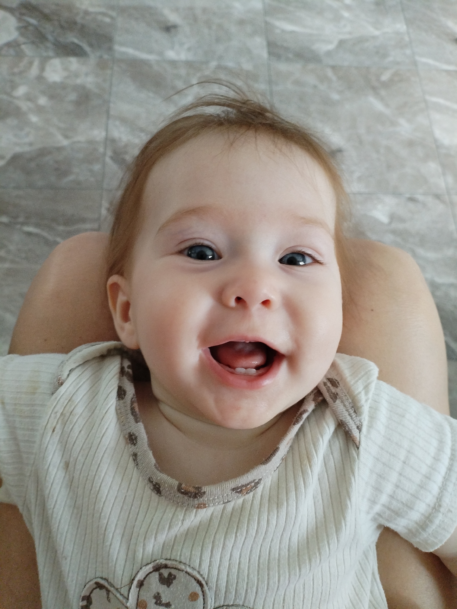 An overhead, eye-level shot captures a Caucasian baby with light brown hair, blue eyes, and a wide, joyful smile with visible teeth. The baby is positioned on someone's lap, indicated by exposed thighs in the foreground. The baby wears a light-colored, ribbed onesie with a patterned trim around the neckline. The background consists of a grey, tiled floor. The scene is indoors. The city of Vinogradny, Russia is not visible in this image.