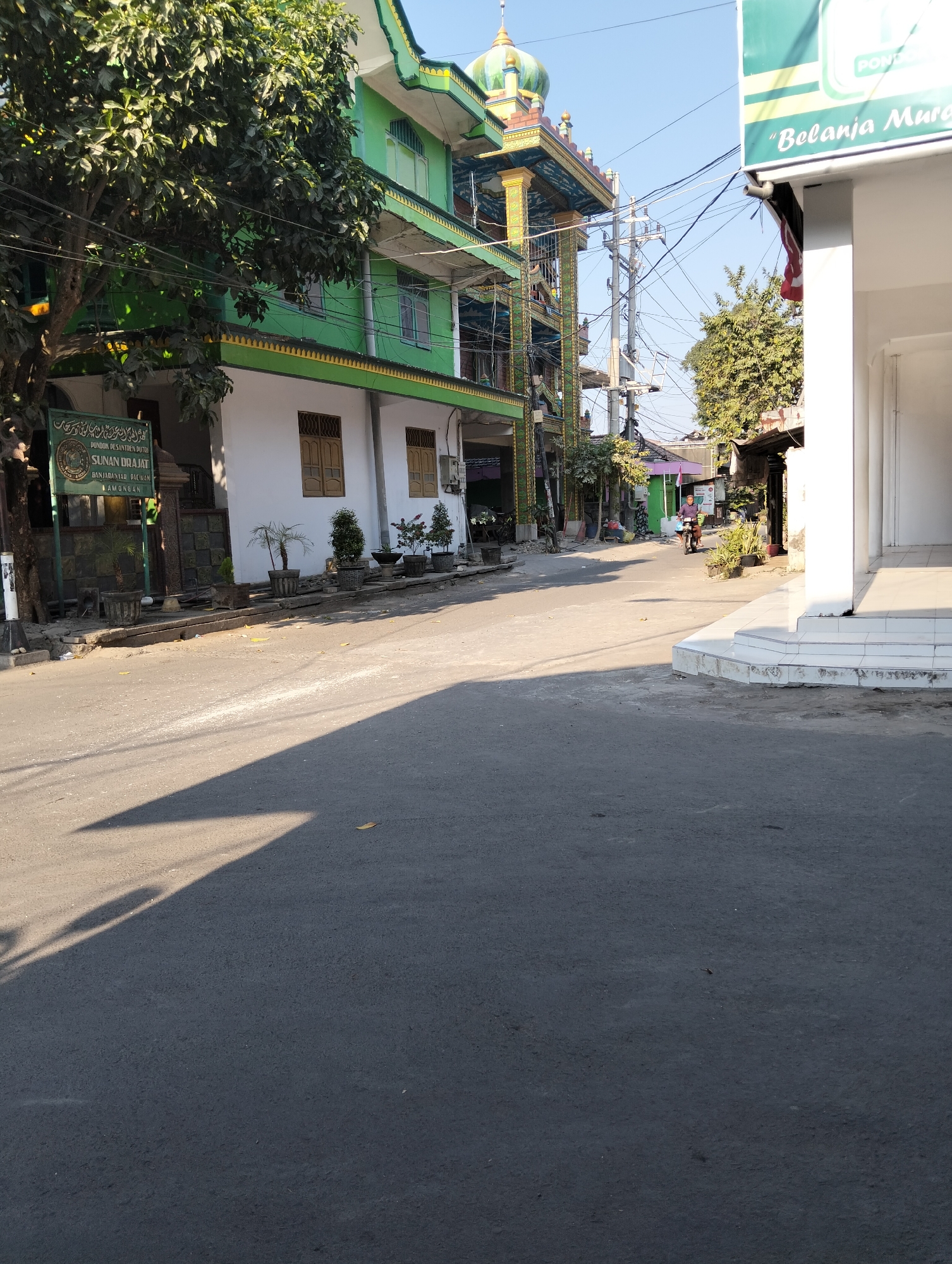 A street scene in Sambipondok, Indonesia, shows a road with a large shadow cast on it from the left. On the left side of the road is a building with green and white walls, featuring a prominent sign for "PONDOK PESANTREN PUTRO SUNAN DRAJAT" and a large tree in front. The building has several windows and a decorative facade. Further down the street, a multi-story building with a mosque-like dome and intricate architectural details is visible. Electrical wires crisscross the sky above. On the right side of the road, a white building with pillars and steps is partially visible. In the background, more buildings and trees can be seen, along with a person riding a motorcycle. The sky is clear and blue.