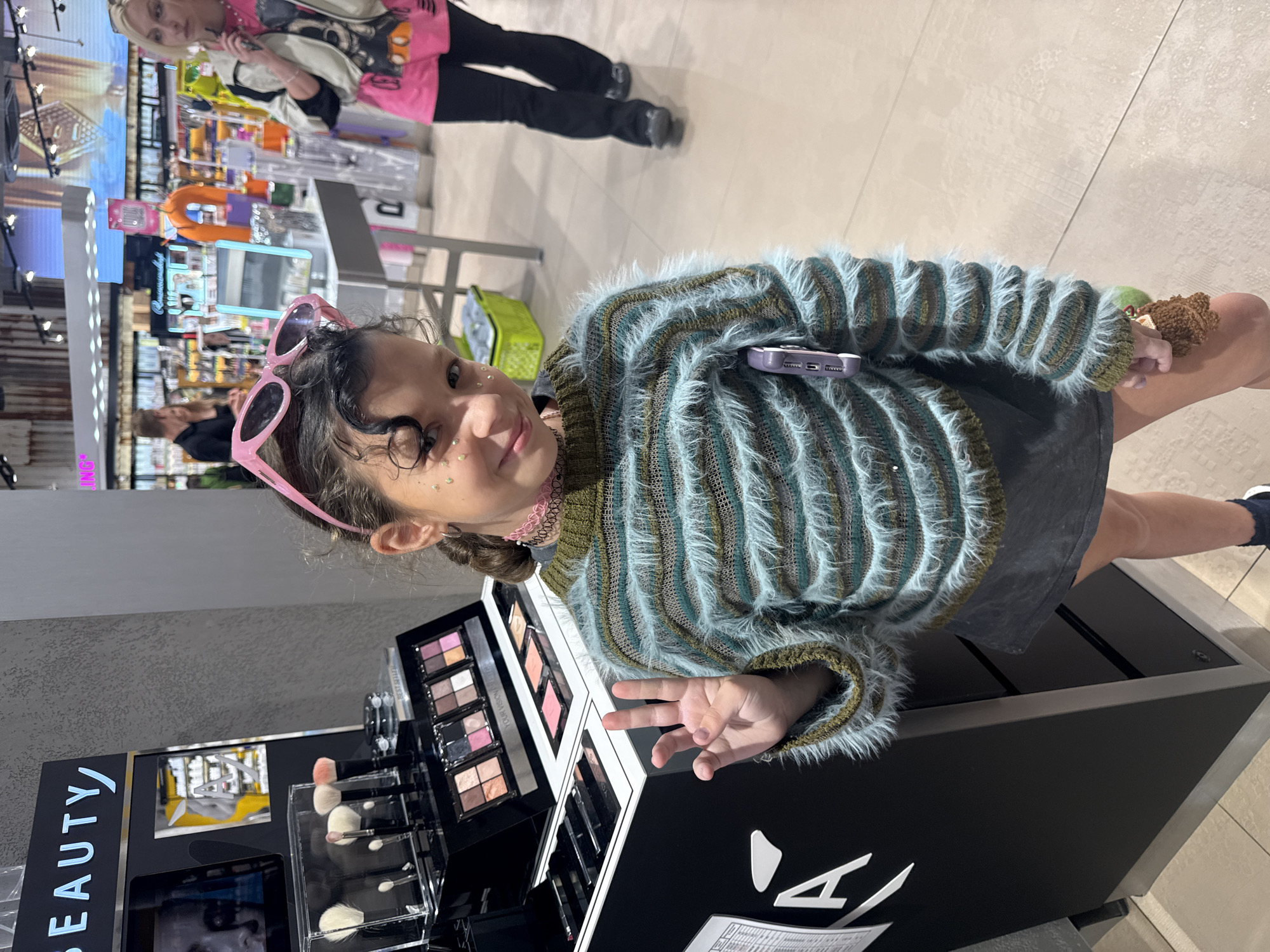 A young girl with glitter on her face and pink sunglasses perched on her head is posing for a photo in a cosmetics store. She is wearing a fuzzy, striped sweater and a dark skirt. Her left hand is raised in a peace sign. In the background, a woman in a bright pink shirt and black pants walks away from the camera. The store appears to be well-lit and stocked with various makeup products. The scene is likely in Saint Petersburg, Russia, given the context of the image.