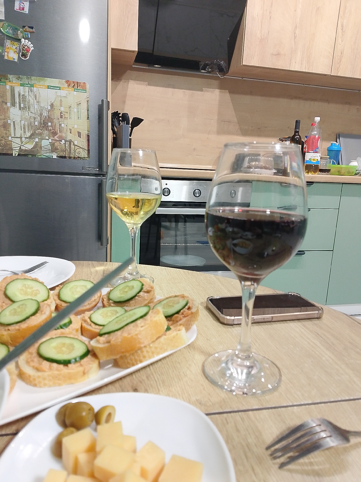 The image shows a table laden with food and drinks, likely in a domestic setting. Two wine glasses are prominent: one filled with white wine, the other with red. A plate of canapés, topped with a spread and sliced cucumbers, is visible. Another plate holds cubes of cheese and olives. A smartphone lies on the table. A fork is placed near the edge of the table. In the background, a kitchen is visible, including a refrigerator with a calendar attached, cabinets, an oven, and assorted bottles and containers. The location, Izhevsk, Russia, cannot be confirmed from the visual information.