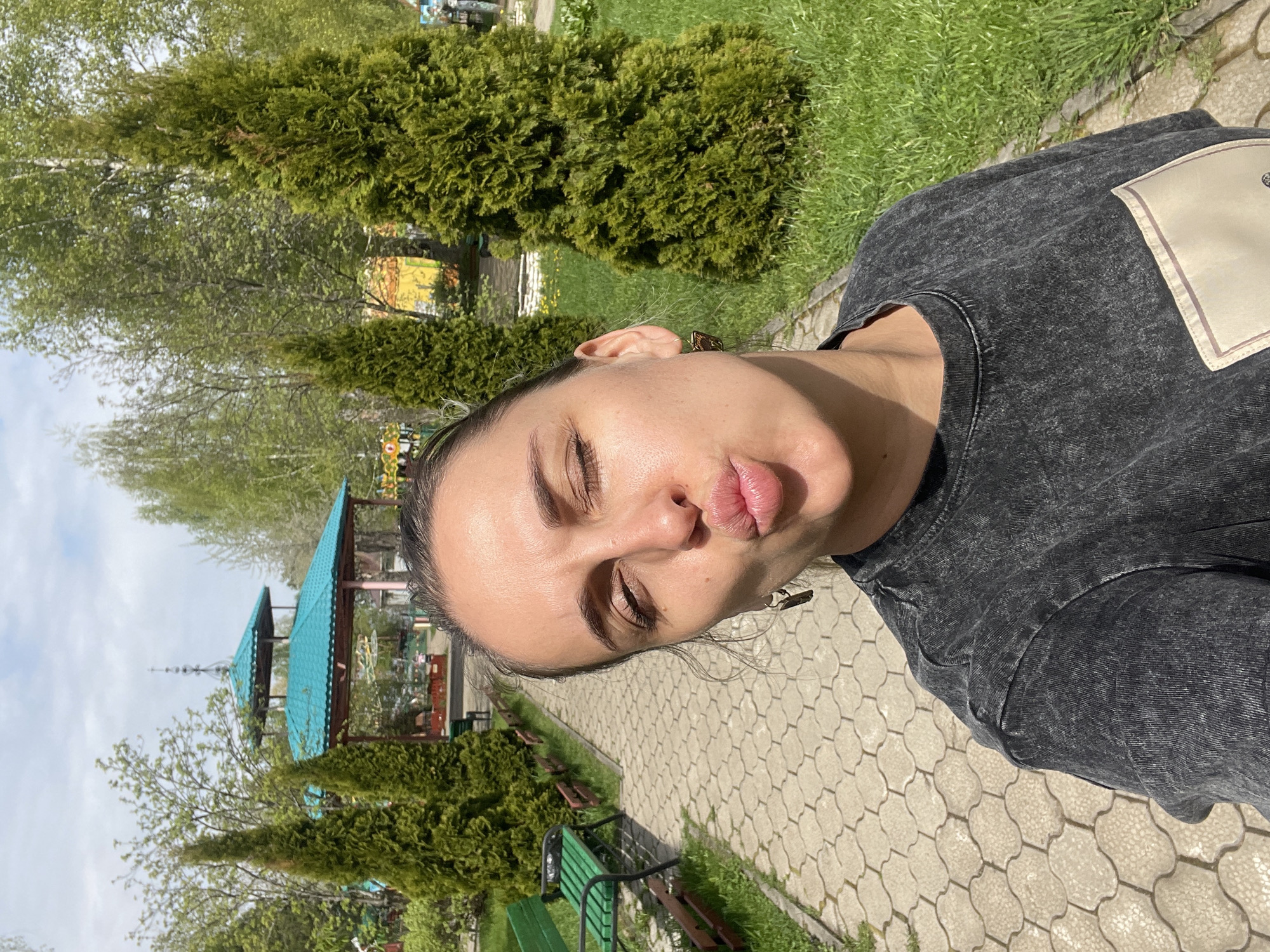 A woman with brown hair pulled back, wearing a dark gray T-shirt with a light-colored patch, is taking a selfie in a park. Her eyes are closed and her lips are pursed. The park is located in Chaikovsky, Russia, as evidenced by the architecture and foliage typical of the region. A stone-paved walkway is visible, lined with green benches, small evergreen trees, and grass. Further back, structures with teal-colored roofs are seen, along with deciduous trees in early spring foliage. The lighting indicates it is daytime.