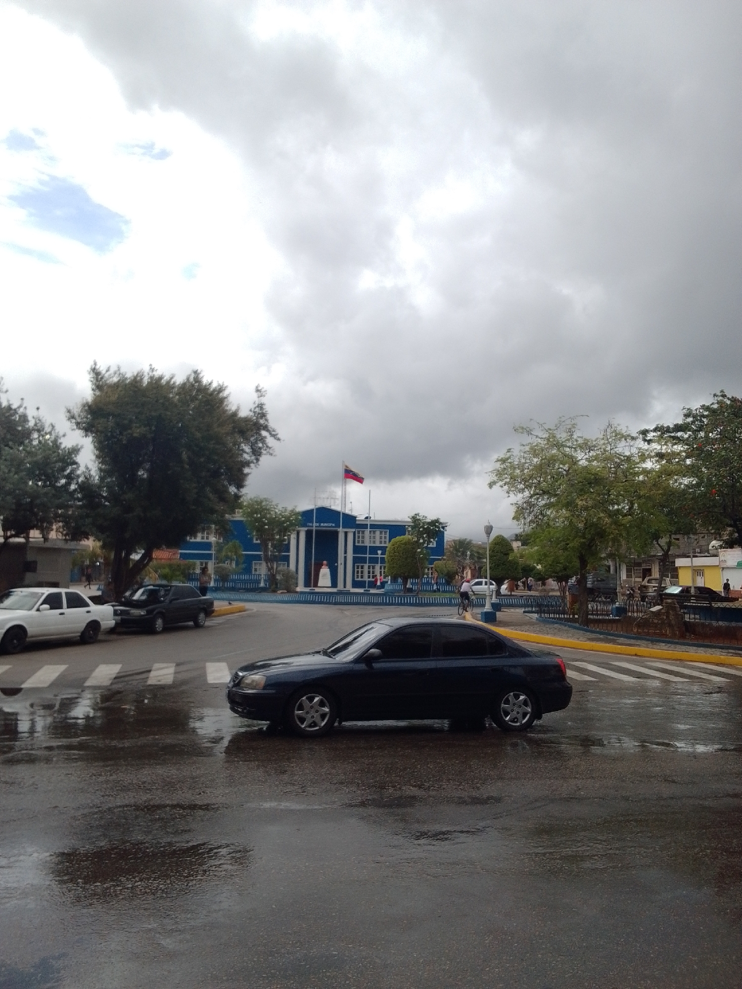 This image captures an outdoor scene in Juan Griego, Venezuela, likely during the daytime under overcast skies. The ground appears wet, suggesting recent rain or ongoing dampness.
The primary subjects include several vehicles. A dark-colored sedan is prominently featured in the foreground, driving on a wet road marked with a crosswalk. To the left, a white sedan and a dark-colored older model car are parked. In the background, a blue municipal building stands with a Venezuelan flag flying from its roof. The building has white columns and decorative blue and white trim.
There are a few people visible, though none are clearly defined. Some figures can be seen near the building and on the sidewalks. A cyclist is also present, riding past the building.
The setting is a town square or a main street area, with trees and landscaping surrounding the municipal building. The overall atmosphere is subdued, possibly due to the cloudy weather. The wet pavement and the grey sky suggest a humid or rainy day. The presence of the municipal building and the flag indicates a public space within a Venezuelan town.
Visible text is limited to "PALACIO MUNICIPAL" above the entrance of the blue building.