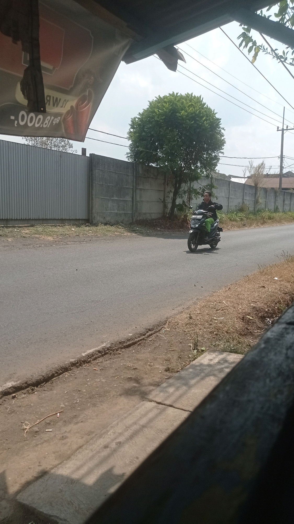 The image depicts a street scene in Bedali, Indonesia, under a cloudy sky. In the foreground, a portion of a wooden structure casts a shadow across a concrete surface with some scattered debris, including cigarette butts. The street itself is paved and appears to be a secondary road.
To the left of the street, a corrugated metal fence runs along the roadside, with a concrete wall behind it. A banner, partially visible, hangs from an overhang above the corrugated fence, displaying text that appears to be in reverse and includes numbers and currency symbols. A lush, rounded tree stands behind the concrete wall, to the right of the banner.
Further down the street, a person is riding a black motorcycle. The rider, wearing a dark jacket and green pants, is looking forward and appears to be moving away from the camera. The motorcycle has a license plate, though the characters are illegible. In the background, more concrete walls and some buildings are visible, along with utility poles and power lines. The general environment suggests a rural or semi-urban area.