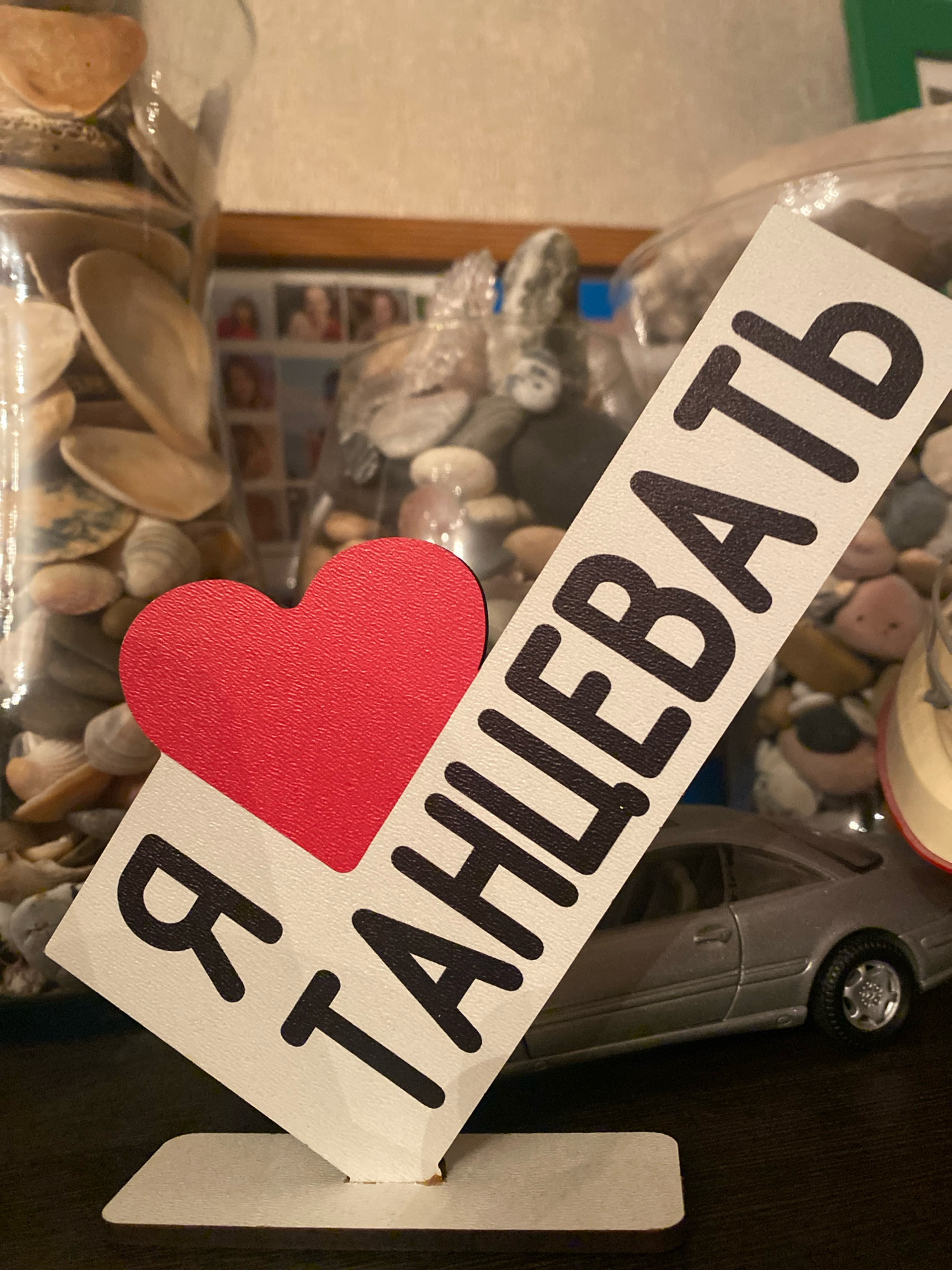 The image shows a close-up of a decorative object displaying a red heart and text that reads "Я ТАНЦЕВАТЬ" (I Dance) in black, stylized Cyrillic lettering on a white diagonal strip. The object is supported by a white base. In the background, there are glass jars filled with seashells and small stones. Behind the jars, a small framed collection of photographs is visible, depicting multiple people. A toy silver car is positioned to the right of the decorative object. The setting appears to be indoors, likely a home or a display area. There is no visible indication of the specific location being in Ivanteyevka, Russia.