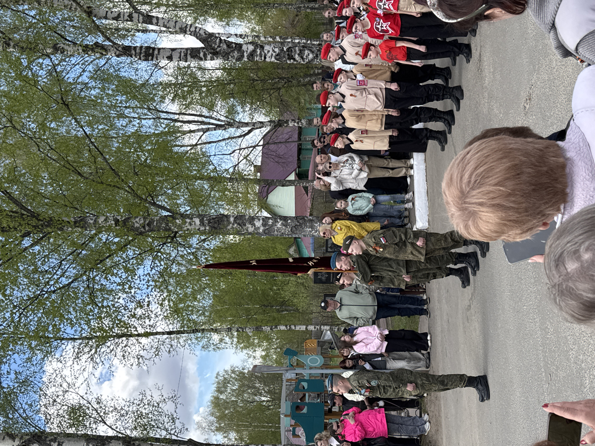 The image shows a scene in the city of Chuprino, Russia. A group of people is standing in rows on an asphalt surface, likely a road or paved area. On one side are individuals dressed in military uniforms, and they appear to be standing at attention, holding a flag. Further down the line are children dressed in red berets and outfits with red star emblems, possibly participating in a ceremony or event. There are also civilian observers in the background. Trees line the edge of the area, with a mix of bare branches and budding green leaves indicating it is springtime. A small building or structure is visible behind the group. The overall impression is of a public gathering, possibly a commemorative event involving military personnel and young people.
