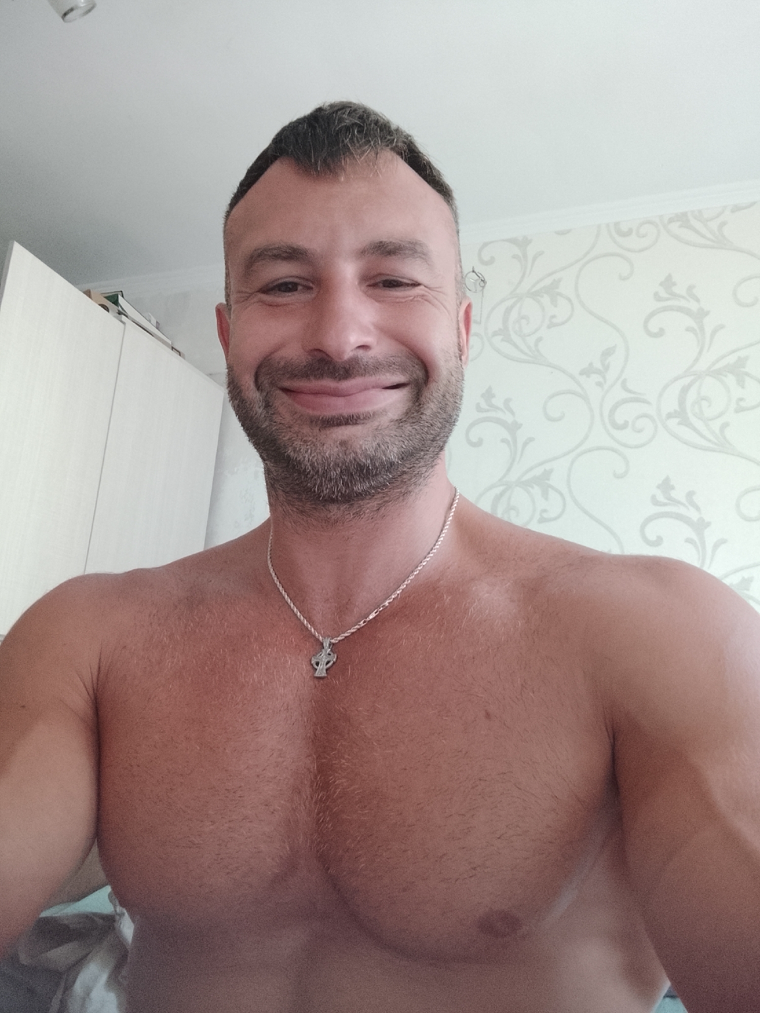 The image is a chest-up selfie of a shirtless man with short, dark hair and a beard. He is smiling and wearing a silver chain with a cross pendant. He appears to be indoors, with a white wall with a floral pattern and a closet visible in the background. There is no information in the image to indicate it was taken in Chișinău, Moldova.