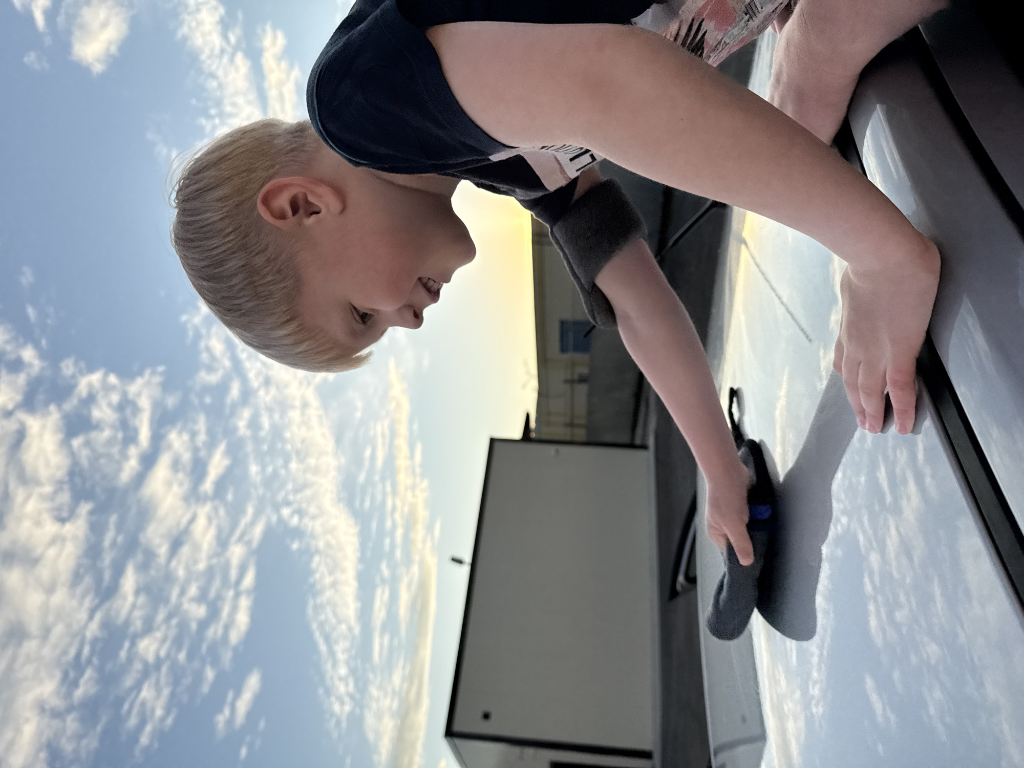 In the city of Chaikovsky, Russia, a young blond-haired boy is observed from a low-angle perspective, leaning over a light-colored car and actively wiping its surface. He has fair skin and short, light blond hair with bangs, and is wearing a dark (likely black or navy) short-sleeved t-shirt with a partial light-colored graphic design visible on his side. A patterned garment, possibly shorts with pink, orange, and white tones, is visible on his lower right side.
The boy's right hand, encased in a dark gray microfiber cleaning mitt with a small blue accent, is scrubbing the reflective silver or light gray surface of what appears to be the car's hood or roof. His left hand rests gently on the car's surface to his left. The car's highly reflective surface mirrors the overhead sky and clouds. The boy's face is turned slightly downward and to the left, showing a gentle smile, indicative of engagement or enjoyment.
The scene is set outdoors under a clear blue sky, sparsely dotted with scattered white, patchy cirrocumulus or altocumulus clouds. The ambient lighting suggests either early morning or late afternoon, with a warm, golden glow visible near the horizon on the right side of the frame. In the background, to the left, stands a modern-looking, rectangular, single-story building with light beige or off-white walls and prominent black trim along its flat roofline and corners. A small, dark, vertical fixture protrudes from its roof. Further to the right in the background, a darker, possibly industrial building is partially obscured, with a fence line visible behind it. The ground appears to be paved, likely asphalt or concrete.