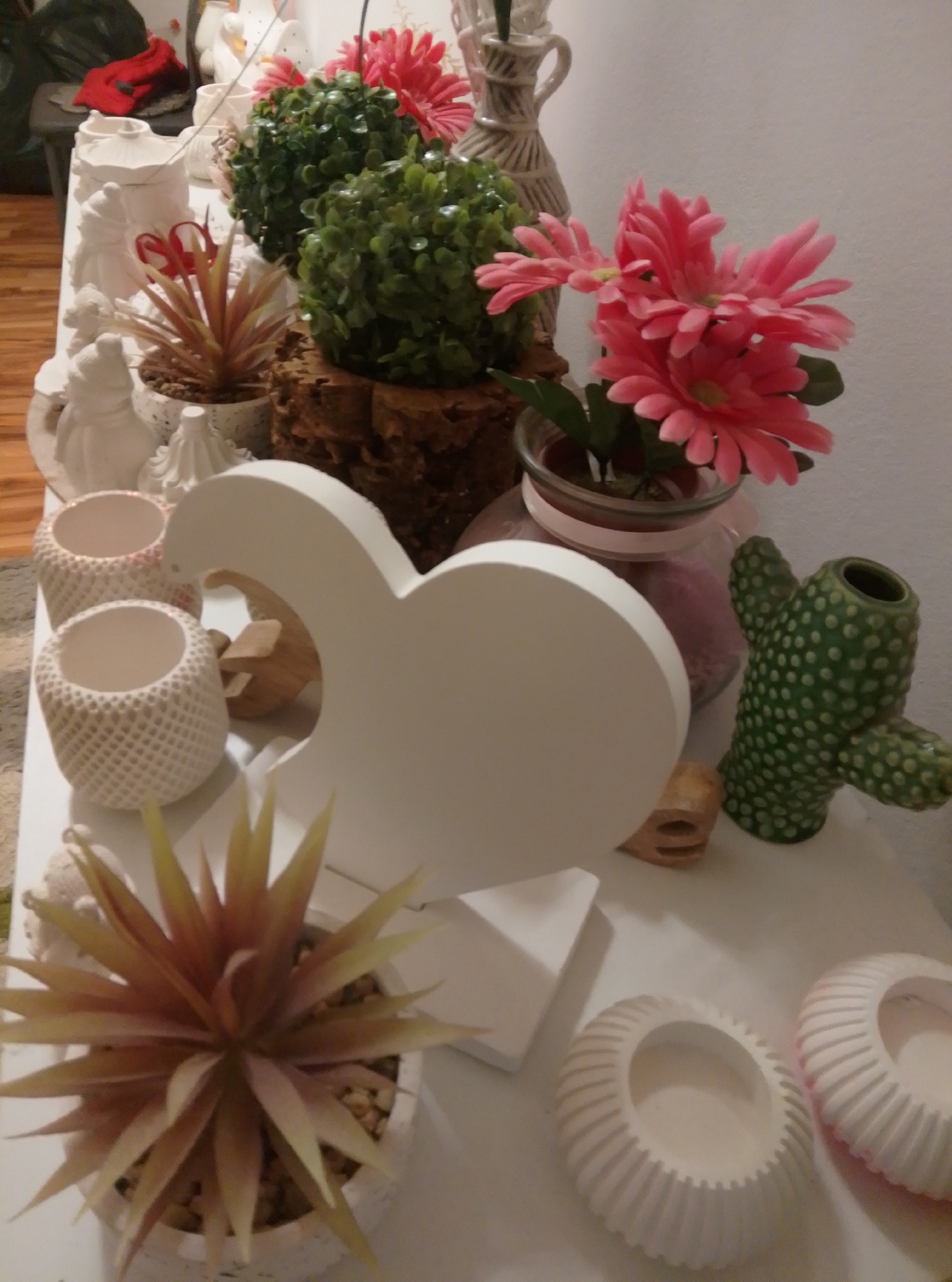 The image depicts a collection of decorative items arranged on a white surface, likely a shelf or table. The items include various artificial plants, ceramic figurines, and decorative pots. In the foreground, a spiky, orange and yellow artificial succulent plant is prominent in a white patterned pot. To its left are two smaller white, patterned cylindrical containers. Behind these, a large, matte white heart-shaped cutout stands upright. Further back, a green ceramic cactus-shaped vase is visible, as well as a glass jar containing pink material and pink artificial flowers. Two spherical, green artificial plants in brown textured pots are also present. A light-colored, ornate vase is situated behind the pink flowers. On the far left, several white ceramic figurines, possibly of people or animals, are arranged, along with a red garment draped over a dark object. The background is a plain white wall. The scene appears to be an indoor display of decorative objects. The image does not provide any information about a specific location in Wedel, Germany, or any specific actions or interactions.