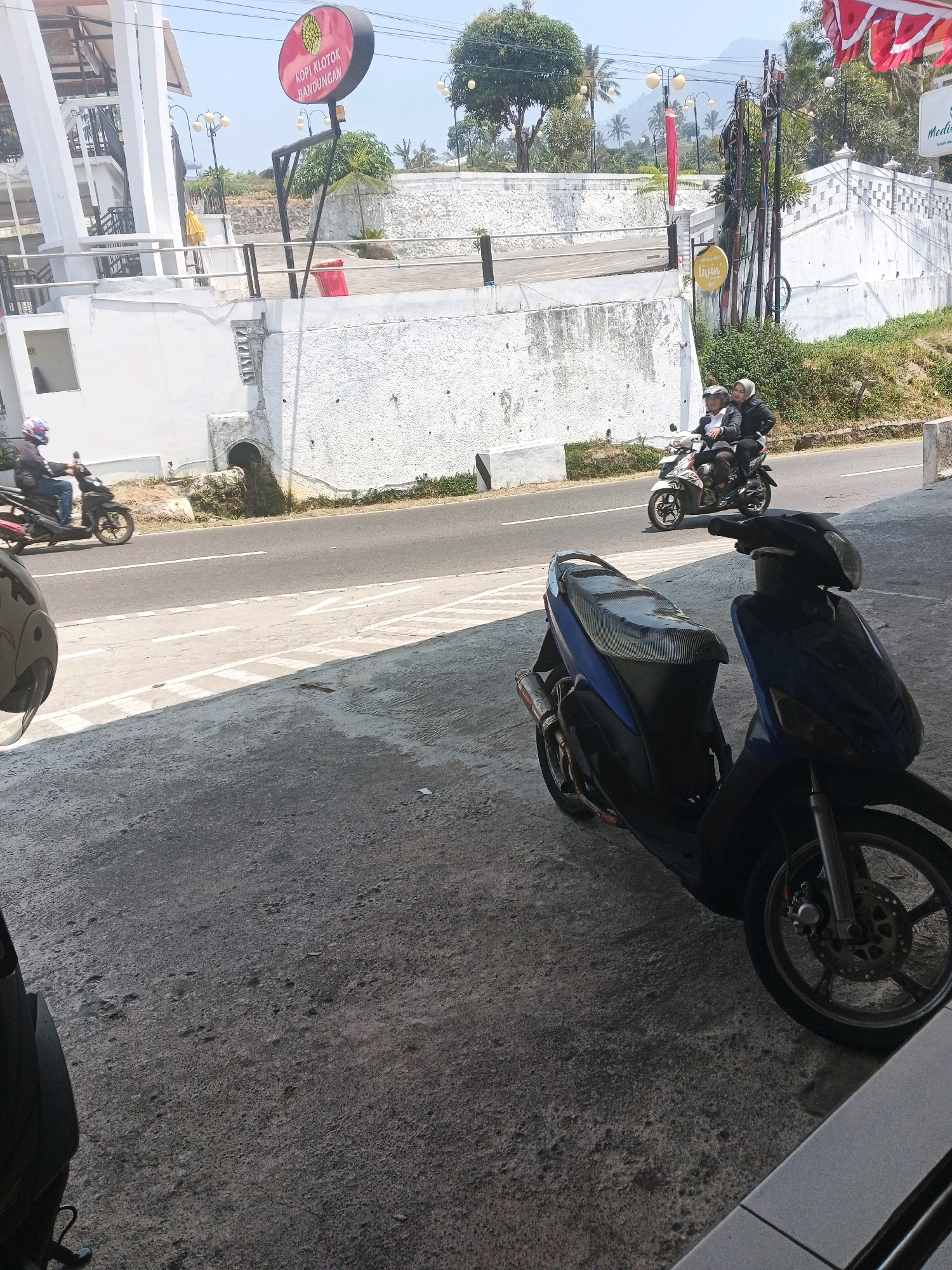 A street scene in Pakopen, Indonesia shows a road with two motorcycles. In the foreground, a dark blue scooter is parked on a paved area. Its seat is covered with a patterned fabric. Further down the road, two individuals on a white motorcycle are riding away from the camera, the passenger is wearing a white hijab. A red trash can is visible near a white building on the left. A circular sign with red lettering on a dark background hangs from a pole above the building, it reads "KOPI KLOTOK BANDUNGAN". The area is illuminated by sunlight, casting shadows on the ground. The background features vegetation and a hazy sky.
