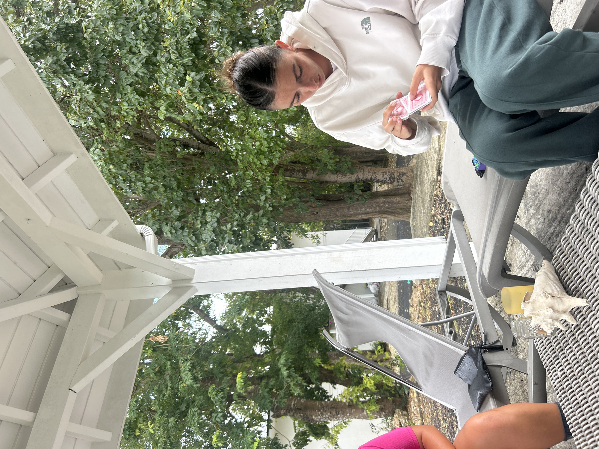 A woman sits on a gray chair on a patio in Sainte-Anne, France. She wears a white hooded sweatshirt and green sweatpants, and is holding a deck of playing cards. The patio is partially covered by a white structure. Outside the patio area are green trees and foliage. A second person is partially visible seated in another gray chair. Other objects nearby include a shell, a yellow container, and a small jar.