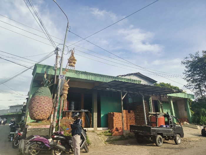 The image shows a street scene in Cikalong, Indonesia, located at coordinates -6.753 latitude and 107.444 longitude. A man in a black helmet and blue shirt stands on a sidewalk near a large, brightly colored pineapple sculpture positioned in front of a building with a green awning and a green facade. The building has a flat roof and an open door and windows. A black pickup truck with a gray tarp-covered canopy is parked on the street in front of the building. The truck has a blue fuel tank on its back, and bricks are stacked in the back of the truck bed. The sidewalk has a few other motorcycles parked on it. The sky is bright blue with white clouds. There are several power lines overhead, and some trees are in the background. The scene is characterized by a combination of urban and rural elements. The presence of the pineapple sculpture and the brick stacks suggest a local market or building supplies shop.