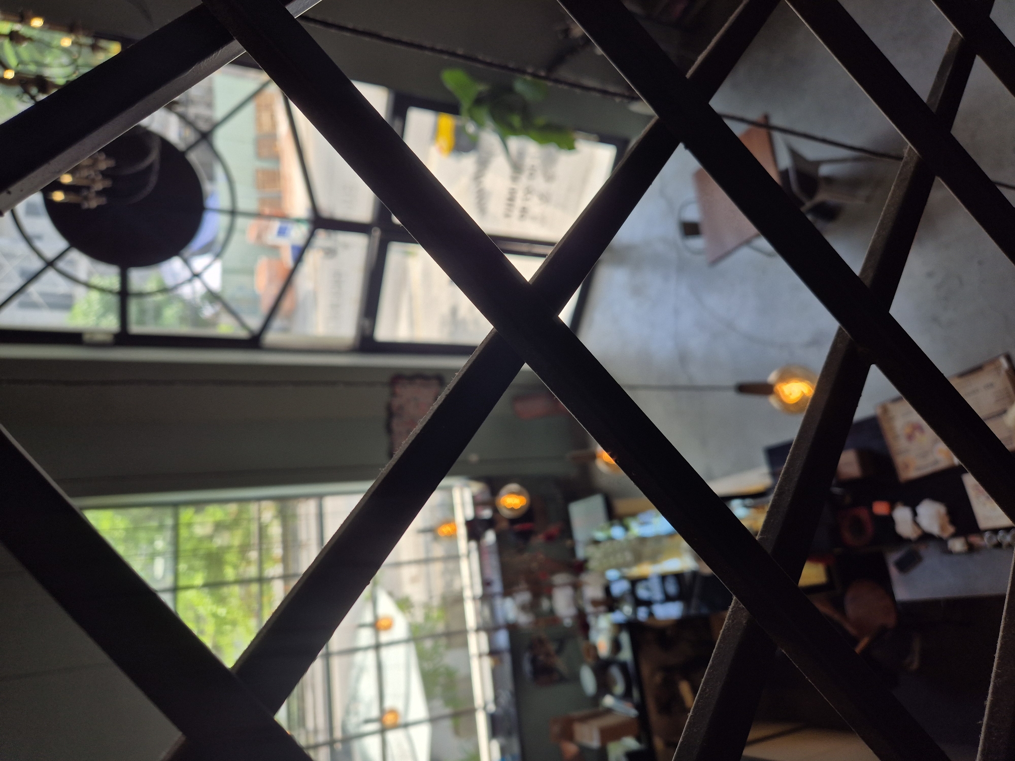 This is a high-angle, indoor shot looking down through a dark, crisscrossing metal grate. The grate creates a diamond pattern that obscures much of the scene below. The environment appears to be a cafe or restaurant with a minimalist, modern aesthetic.
Through the openings of the grate, we can discern several distinct areas. To the left, a large window reveals lush green trees and foliage outside, suggesting a daytime scene with natural light. Below this window, tables and chairs are visible, along with what looks like a counter area with some items on display. Several pendant lights with warm, orange glows hang from the ceiling, contributing to the ambient lighting of the interior.
Further into the space, a prominent large, circular window with a stylized geometric pattern is visible. This window looks out onto a street scene, with buildings and a yellow vehicle visible in the distance. Within the cafe, more tables and chairs are scattered throughout, some with people seated at them, though their features are indistinct due to the distance and the grate. One table in the foreground of this area is a light brown square shape with chairs around it.
The overall impression is of a contemporary urban establishment, likely a cafe or restaurant, possibly located in Đà Nẵng, Vietnam, given the tropical vegetation visible outside the window and the general architectural style. The grate in the foreground adds a layer of visual complexity and depth, framing the view of the interior and exterior spaces.