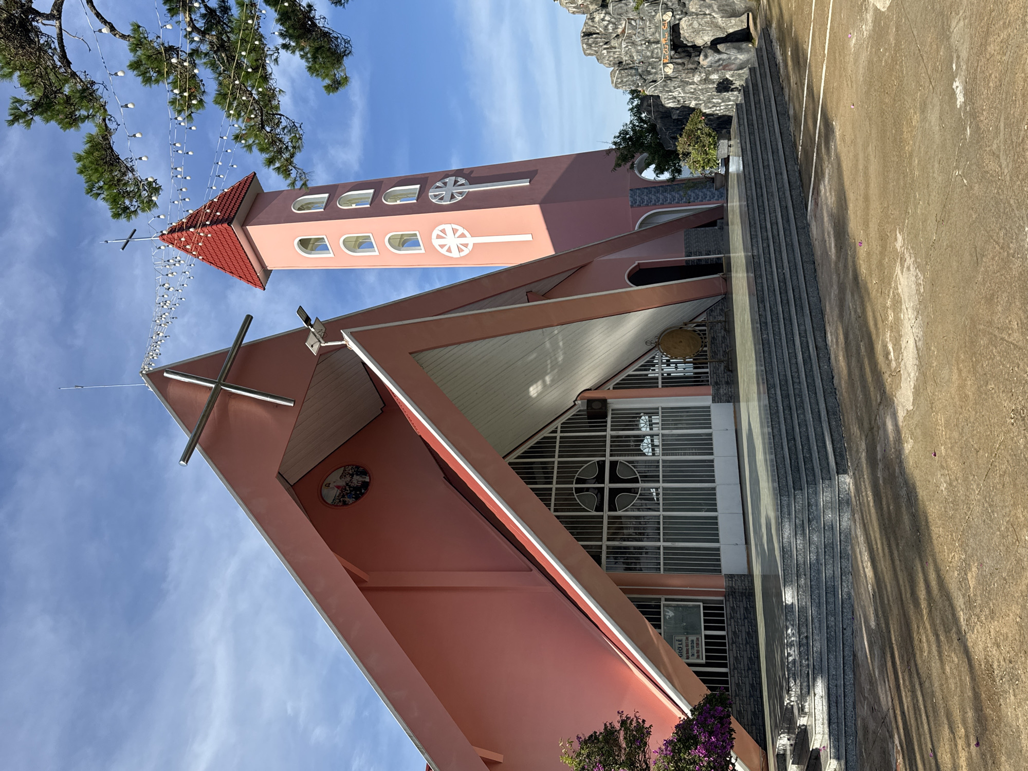The image shows a pink church with a tall, slender tower in Đa Thọ, Vietnam. The church features a distinctive triangular roof with a white trim and a large cross at its peak. The tower, also pink, has a red roof and is adorned with arched windows and a prominent cross symbol. String lights are draped across the scene, suggesting a festive or decorative element. In the foreground, there are wide stone steps leading up to the church entrance, which is comprised of large, metal-framed glass doors. To the right of the church, a rocky formation with carved figures and text is visible. The ground in front of the church is a paved courtyard with some scattered pink flower petals. The sky is a clear blue with some wispy white clouds. The overall scene conveys a bright and serene atmosphere.