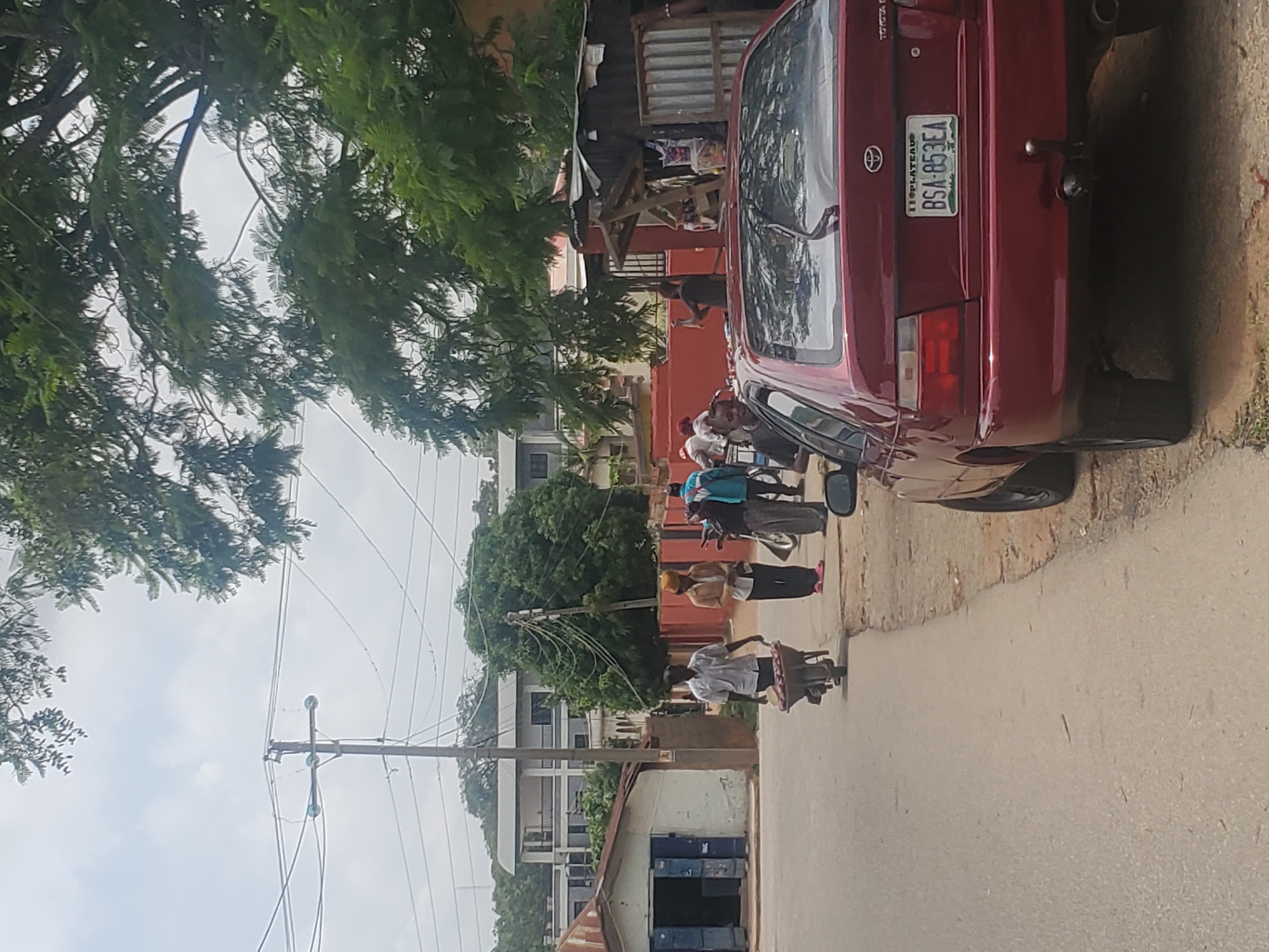 The image depicts a street scene in Jos, Nigeria. In the foreground, a red Toyota car with a license plate reading "BSA-853EA" is parked facing away from the camera. To the left of the car, a paved road curves to the left. Further down the road, several people are walking. One man is pushing a wheelbarrow filled with red produce. Another person, wearing a yellow head covering, stands on the sidewalk. A small, weathered building with blue shutters is visible on the left side of the street. In the background, trees and buildings can be seen. A utility pole with electrical wires is also present. The sky is overcast.
