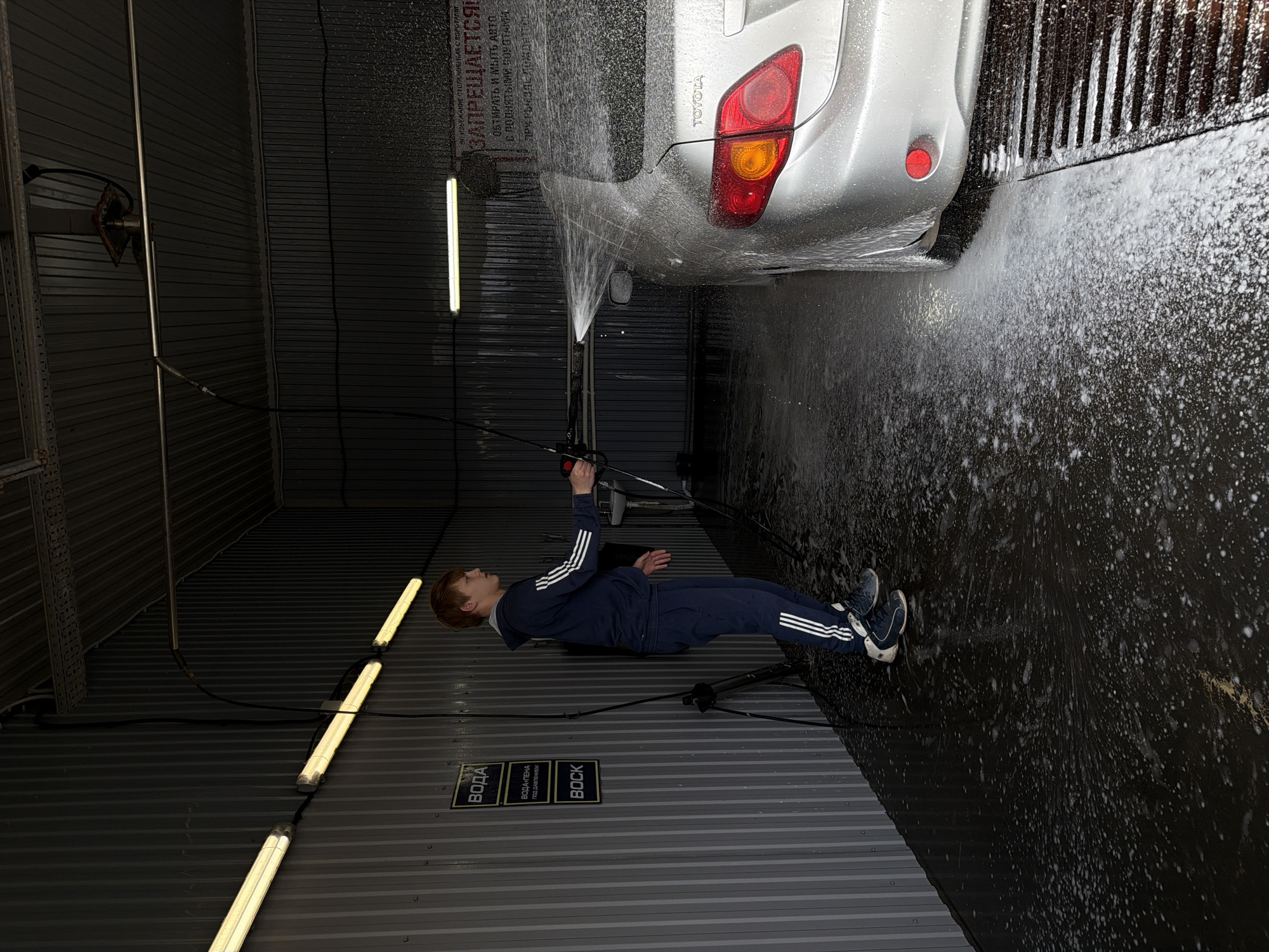 A rotated wide-angle shot shows a man washing a silver Toyota in what appears to be a self-service car wash. The car is positioned with its rear end toward the upper right corner of the image. The man, wearing a blue Adidas tracksuit and sneakers, is holding a pressure washing wand and directing the spray toward the car. The floor is wet and covered in soapy foam. The interior of the car wash is a metallic corrugated structure. Fluorescent lights are mounted along the wall on the left side of the frame. A sign with text in Russian ("ВОДА", "ВОДА ПЕНА ПОД ДАВЛЕНИЕМ", "BOCK") is visible on the wall. Another larger sign with more Russian text is above the car, indicating the location is Chaikovsky, Russia.