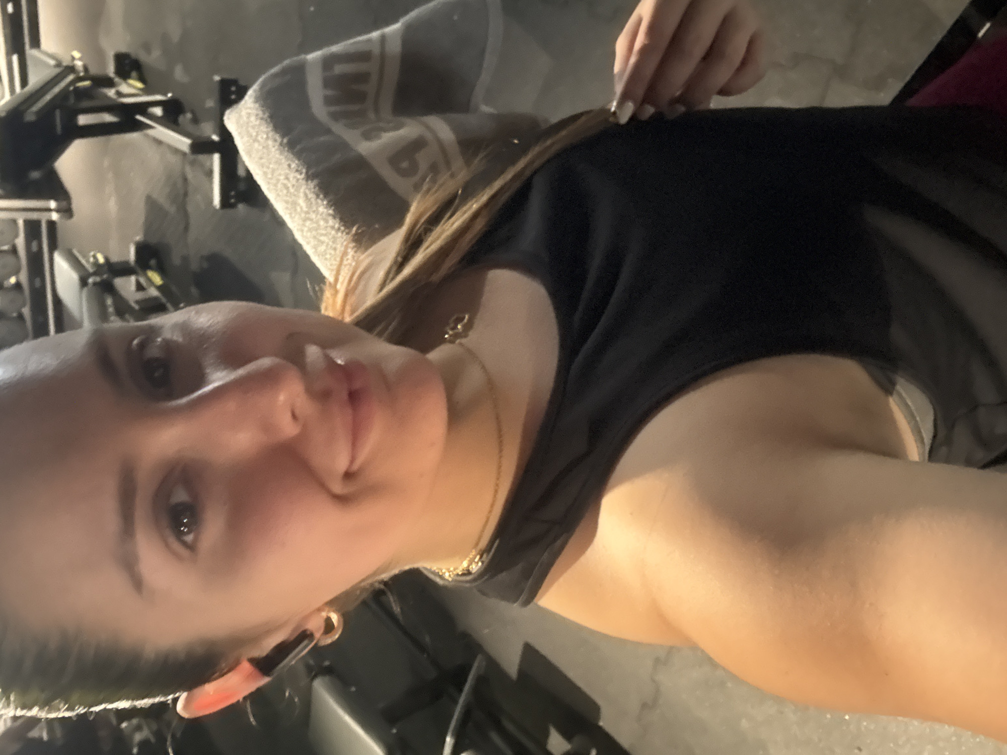 A woman is in a gym setting, with exercise equipment visible in the background. She is wearing a black tank top and a gold necklace and has an earring. A grey towel is draped nearby. She is looking towards the camera. There is no indication that this photo was taken in Barcelona, Spain.