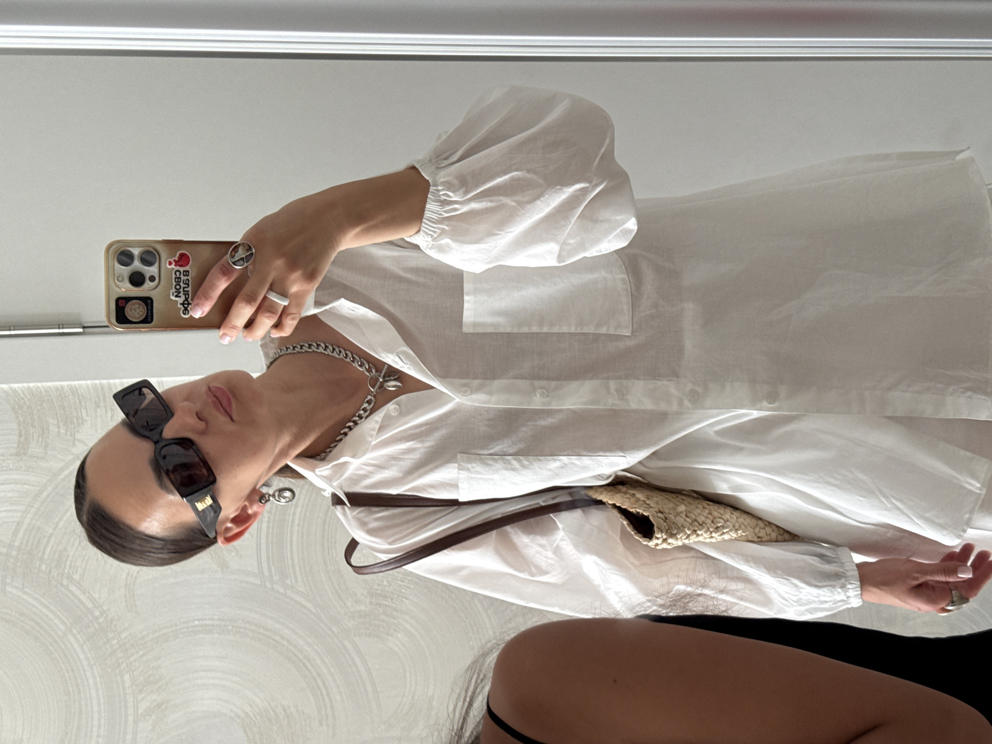A woman takes a mirror selfie in what appears to be a brightly lit, minimalist interior. She is wearing oversized sunglasses with dark lenses, a white button-down shirt with voluminous sleeves, and silver jewelry including a chunky chain necklace, hoop earrings, and rings on multiple fingers. A straw bag with brown leather straps is slung over her shoulder. Her phone, with a gold case adorned with stickers, is visible as she holds it up to take the photo. A reflection of another person, wearing a black tank top, is visible to the left of the frame. The background wall has a textured, wavy pattern. The environment suggests a contemporary setting within a city. There is no indication of the specific location being Sochi, Russia.
