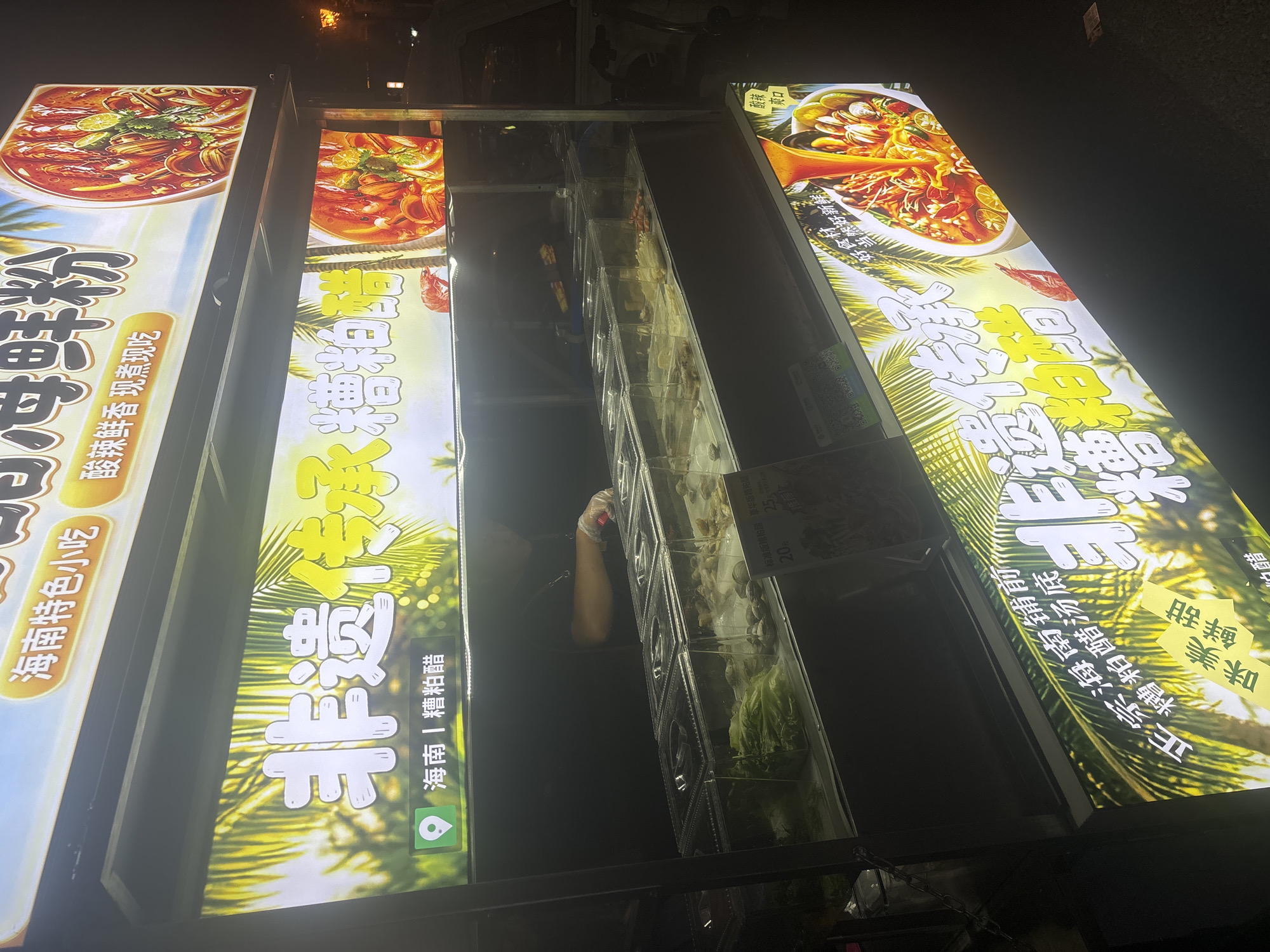 The image shows a brightly lit food stall at night in Suzhou Industrial Park, China. The stall features several large illuminated signs advertising seafood noodles. The signs display images of seafood dishes, Chinese text, and palm trees. Below the signs, transparent containers hold various ingredients, including lettuce and shellfish. A person, likely a vendor, is visible behind the counter, wearing gloves and appearing to prepare food. The stall is positioned on a street, with a vehicle visible in the background. The overall scene is indicative of a street food vendor operating in a commercial area.