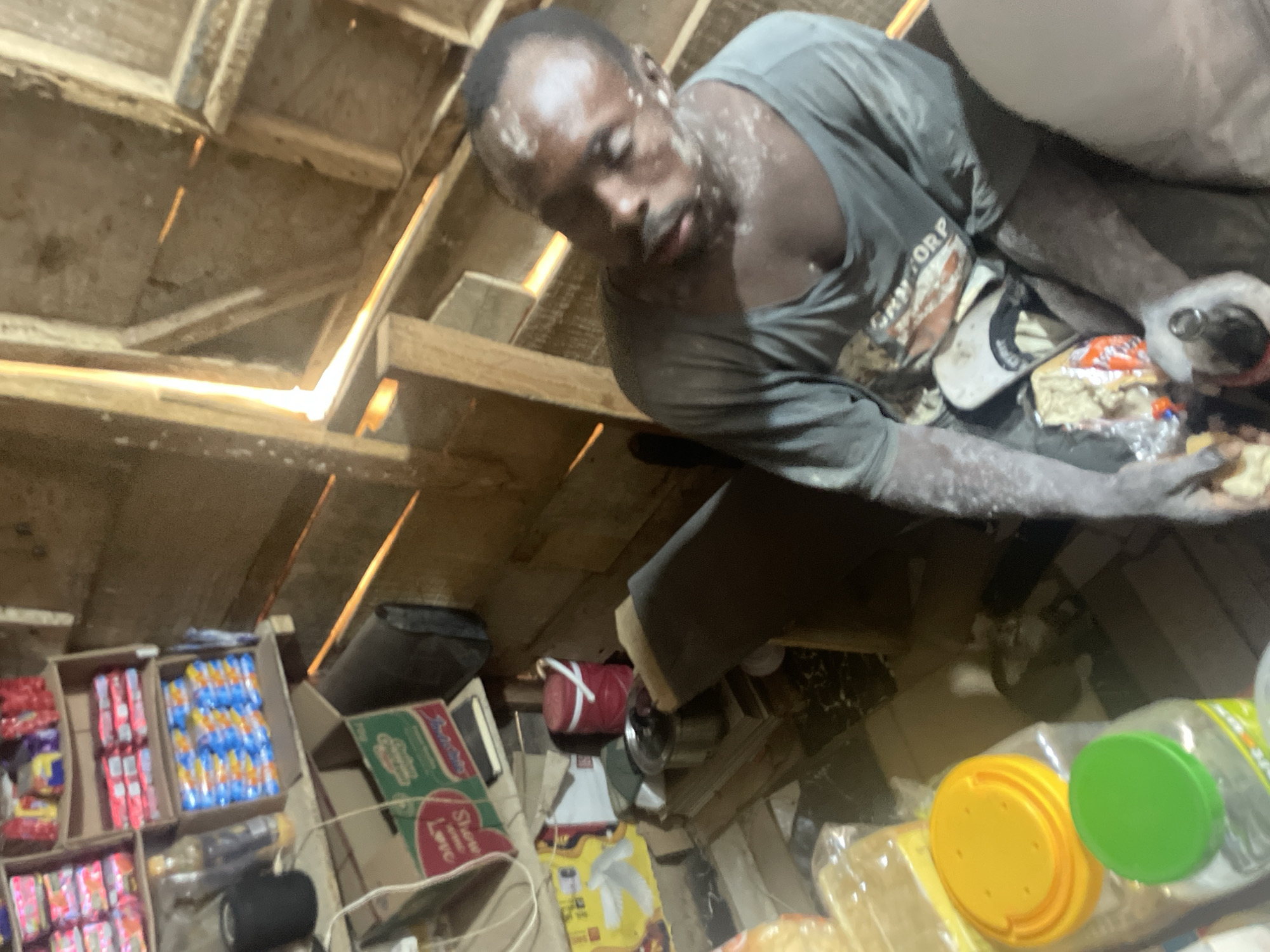 A low-angle shot in what appears to be a small shop or storage space. A dark-skinned man with white powder on his face and shirt is at the top of the frame, looking down and holding something wrapped in plastic. Boxes of goods are visible on shelves along the bottom and left side of the frame. The walls and ceiling are unfinished wood. The location is Abuja, Nigeria.
