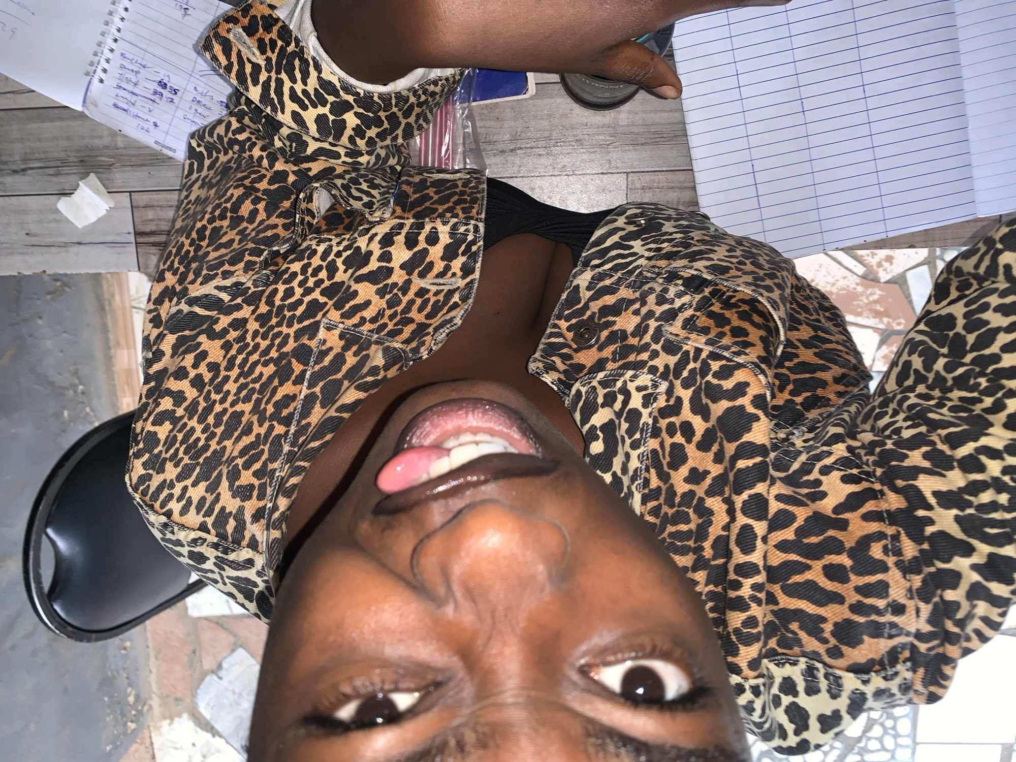 The image shows a close-up, rotated view of a young Black woman wearing a leopard-print jacket and a black top. She is sticking her tongue out, with her mouth open, revealing her teeth. Her eyes are wide open, and she appears to be looking directly at the camera. The background consists of a wooden surface, with a notebook open to a page with blue lines and some writing, as well as a crumpled piece of white paper. A black chair is visible in the upper left corner. The setting appears to be an indoor environment. There is no specific indication of the location being in New Stadium, Nigeria.