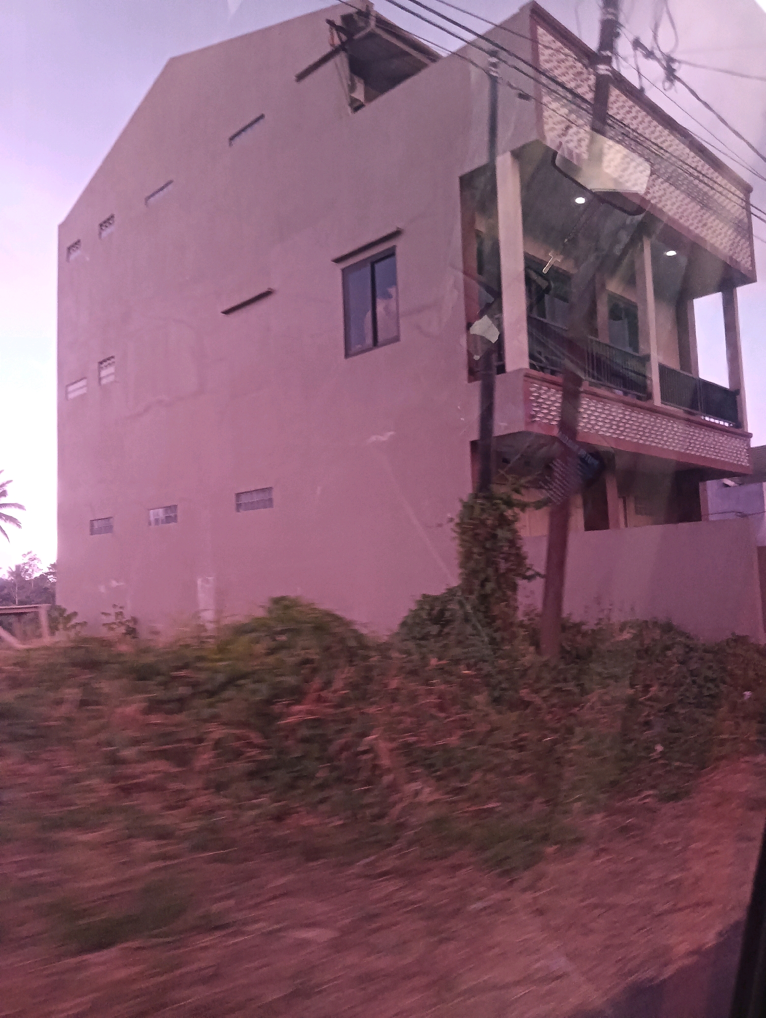 The image shows a tall, narrow, modern-style house with a pale pink facade. The house has multiple stories, with visible windows and balconies. The lower part of the house is partially obscured by dense, overgrown vegetation and a blurred foreground, suggesting the photo was taken from a moving vehicle. Power lines are visible overhead, and a utility pole stands near the house, entwined with vines. The sky above is a pale, hazy purple, indicative of either dawn, dusk, or possibly smog. The overall impression is of a partially built or somewhat neglected urban or semi-urban environment in Tomohon, Indonesia.