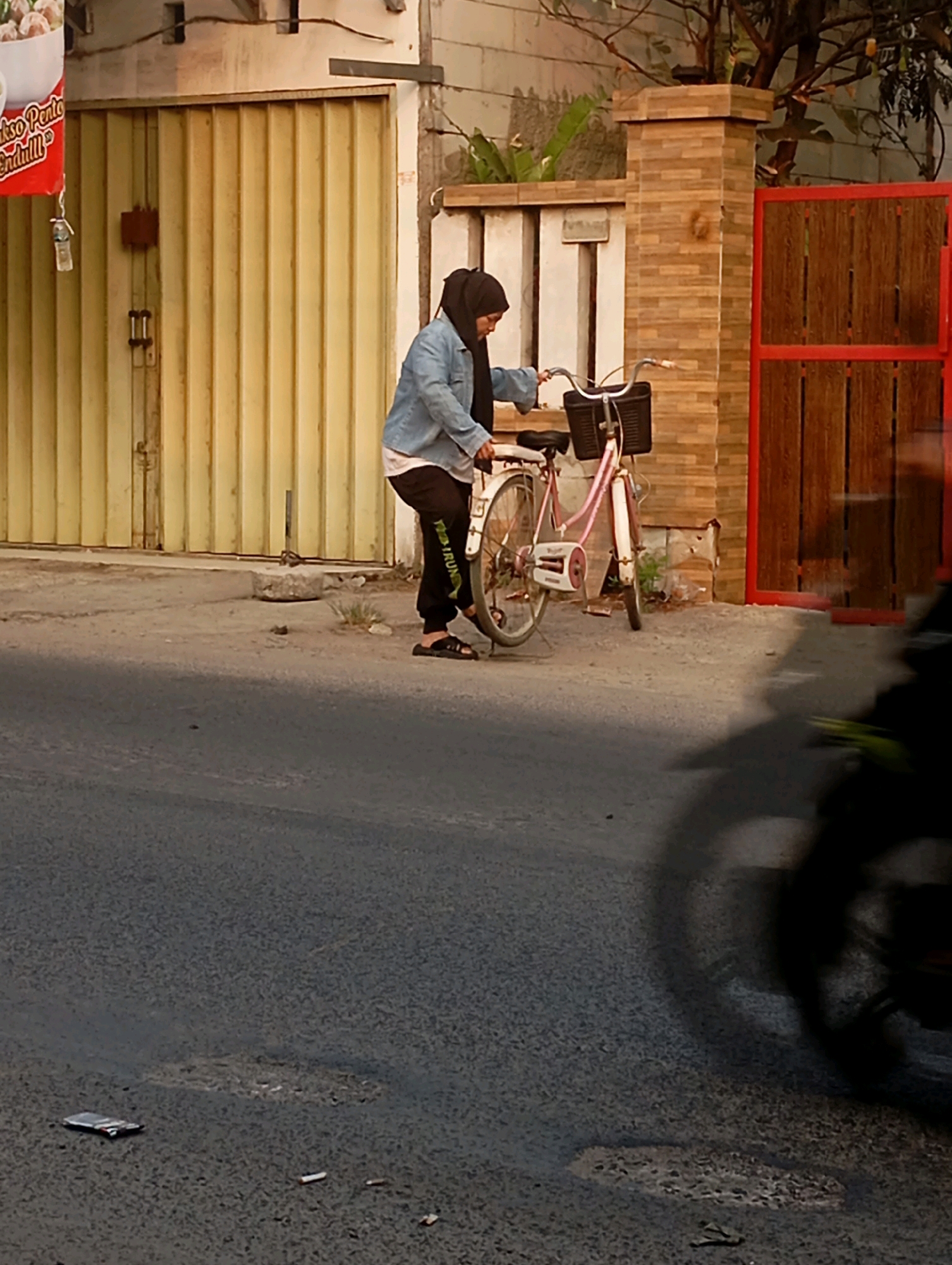 In Babelan, Indonesia, a person wearing a hijab and a denim jacket is standing next to a pink bicycle. The person is looking down at the bicycle and appears to be adjusting it. The bicycle has a basket on the front and a red stripe on the frame. The scene is set in front of a building with a yellow corrugated metal door. A brick fence with a red gate is visible to the right of the building. The ground is paved with asphalt and there is some debris on the road. A blurred image of a motorcycle is in the foreground, casting a shadow on the road.