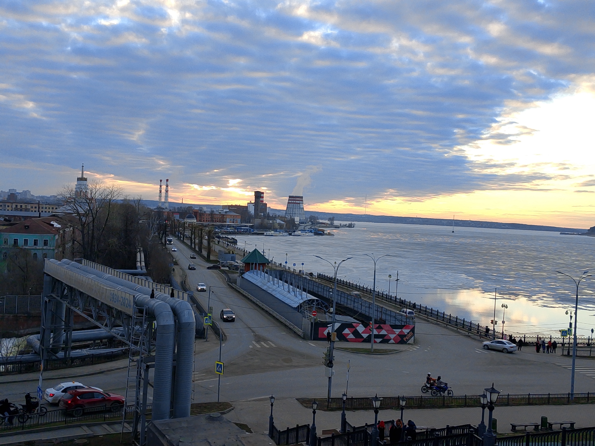 The image depicts a cityscape at dawn or dusk, featuring a wide river with patches of ice. A thermal power plant is visible in the background with tall structures and smoke plumes. The city of Izhevsk, Russia, is suggested by the presence of the IzhAvto heat pipe bridge over the road, and the Votkinsk thermal power plant at the edge of the city, situated between the Izhevsk pond and the Votkinsk reservoir.
A road runs along the riverbank, with occasional cars and pedestrians. There's a pedestrian overpass with piping. A crosswalk is visible at the foreground intersection, with motorcycles waiting and pedestrians in the crosswalk. Street lamps line the embankment. The sky is filled with layered clouds, colored with the warm hues of the setting or rising sun.