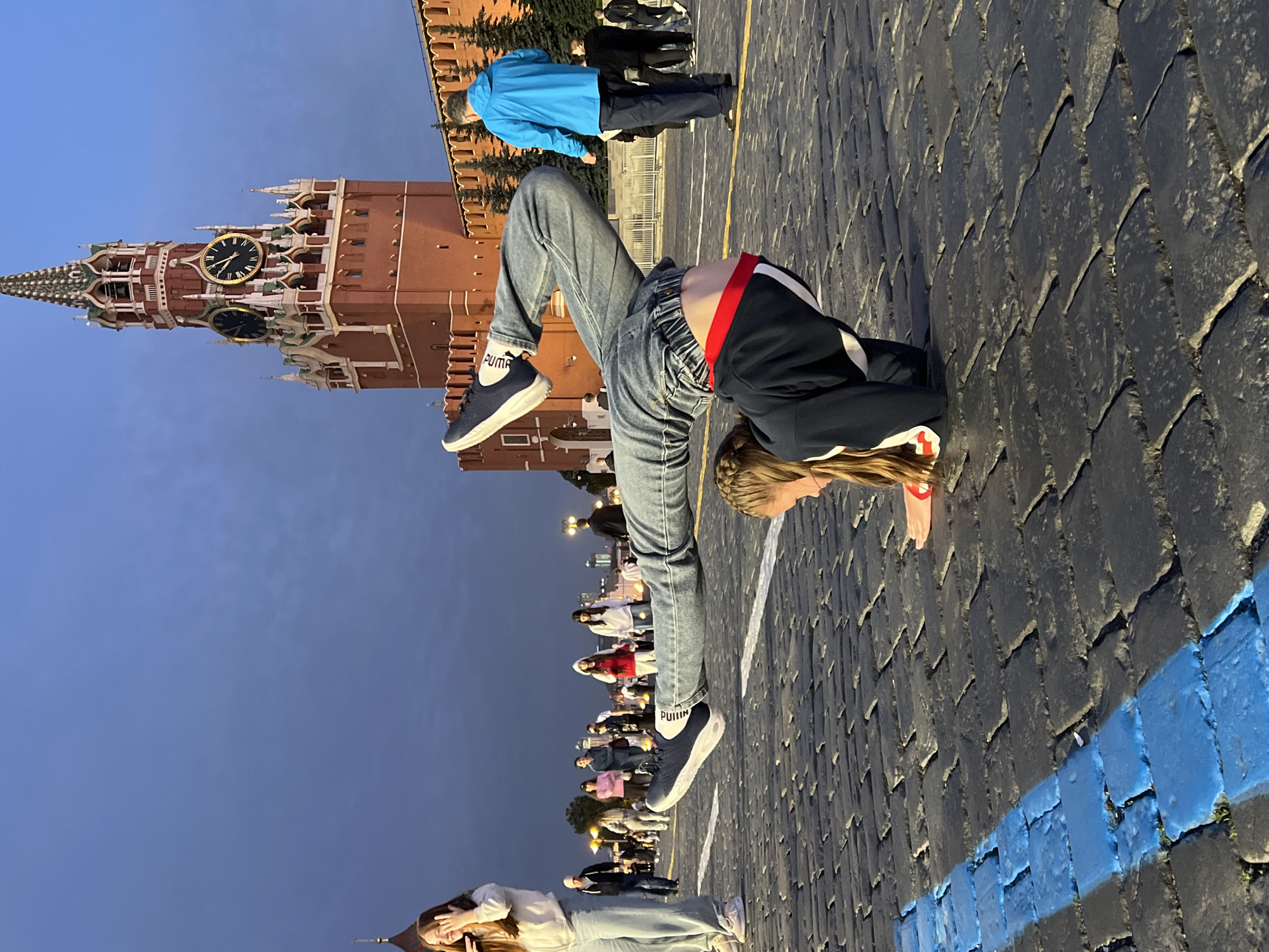 The image shows a group of people on a cobblestone plaza in front of a red brick building with a clock tower. The sky is a deep blue, suggesting it is either dusk or dawn. The building appears to be a historical landmark, possibly a palace or a government building, with intricate architectural details and spires. Several people are standing and walking around, while one person in the foreground is performing a handstand on the cobblestones. This person is wearing jeans, a navy blue jacket with white stripes, and dark sneakers with white socks. The scene is illuminated by artificial lights, indicating it is not full daylight. A bright blue line is painted on the cobblestones in the foreground. The overall atmosphere seems casual and public, with people enjoying the open space.