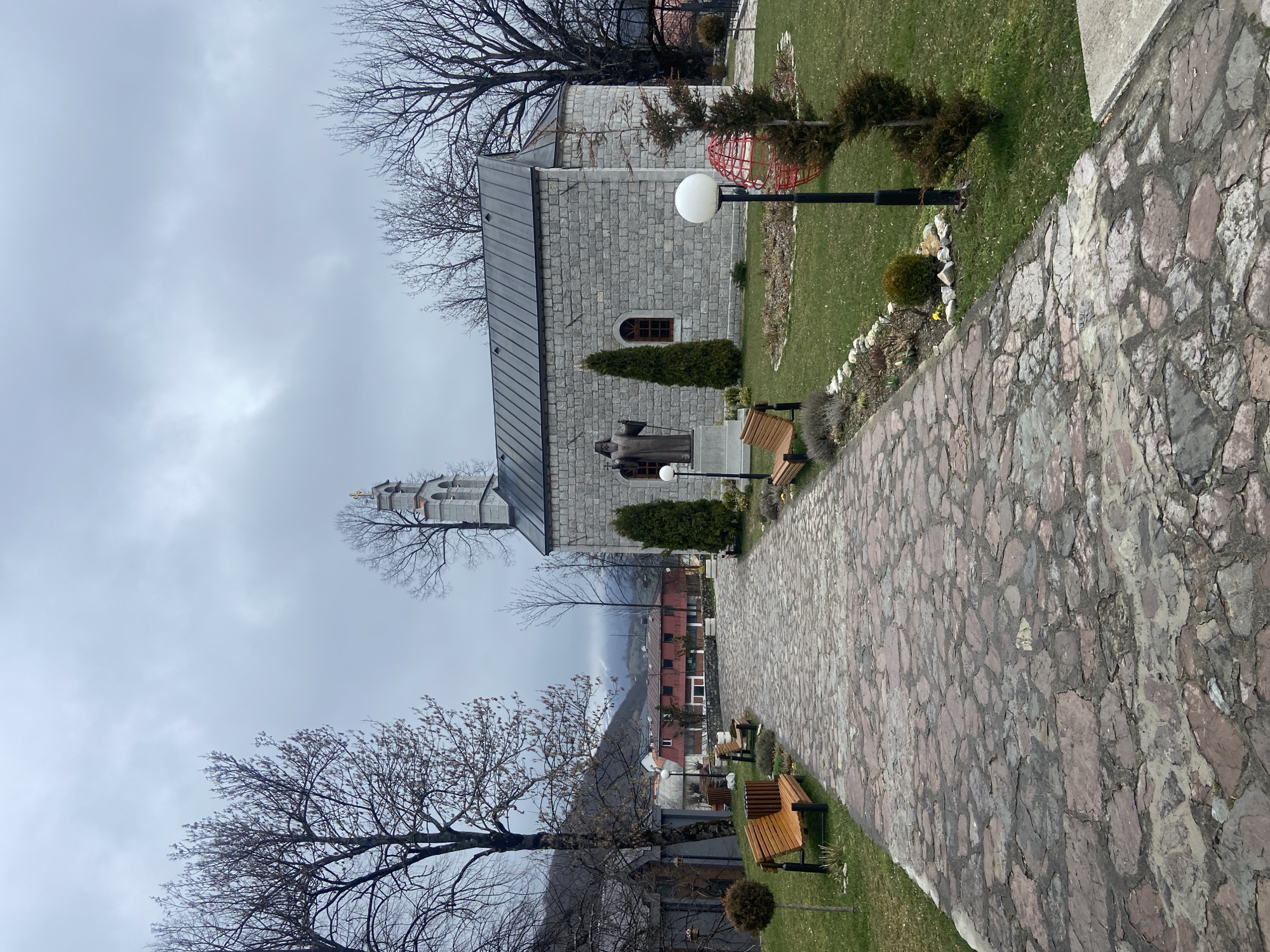 The image shows a stone path leading to a stone building in Kolašin, Montenegro. A statue stands in front of the building. There are bare trees on either side of the path, and benches are placed along it. The sky is overcast.