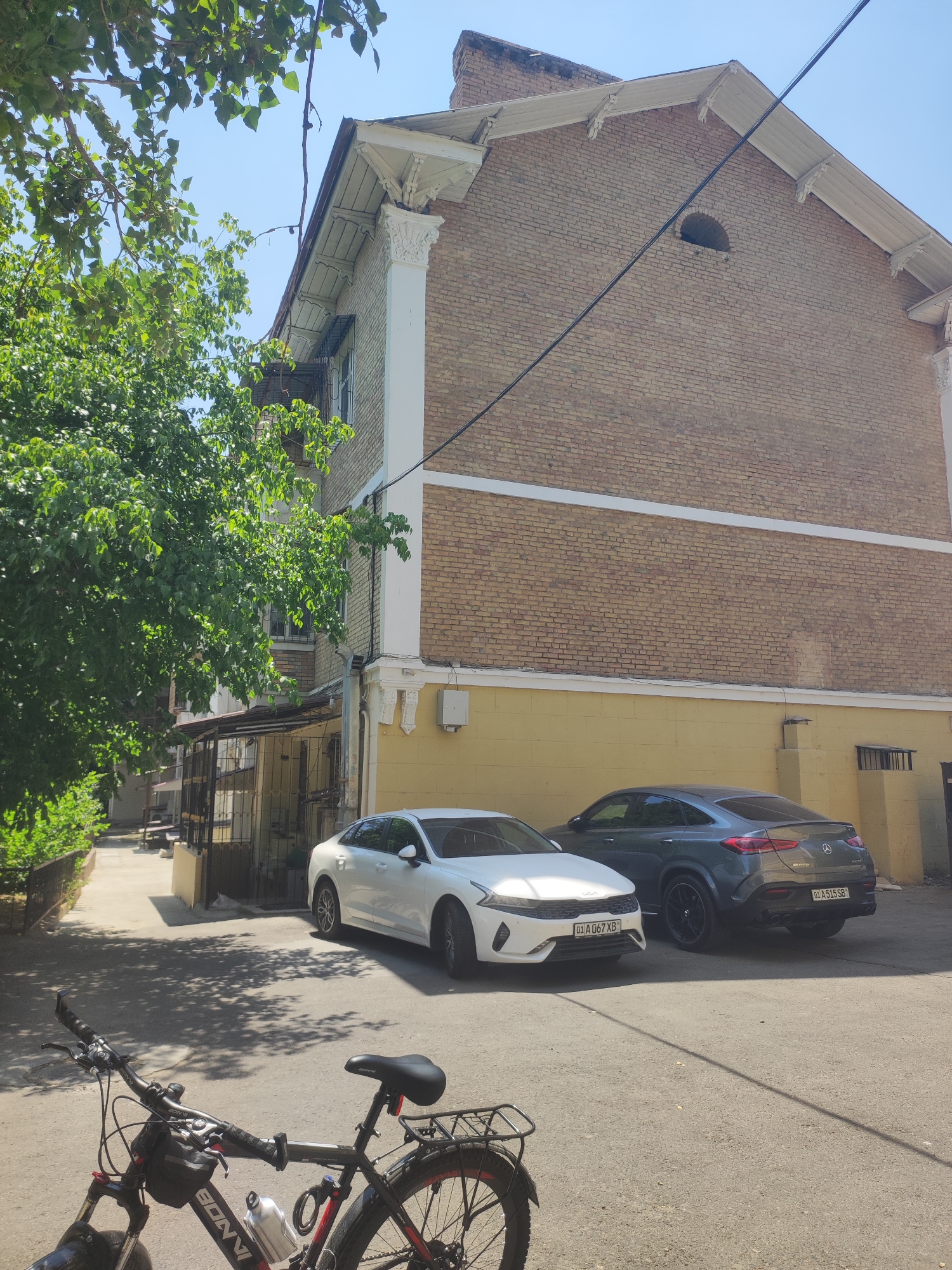 The image shows a sun-drenched street scene in Tashkent, Uzbekistan, with a residential building dominating the background. The building, constructed with yellow brick, features white decorative elements along its roofline and corners. A mature tree with lush green foliage extends from the left side of the frame, casting dappled shadows on the asphalt.
Parked on the street are two modern cars: a white sedan on the left and a dark grey coupe SUV on the right. Both cars have license plates, suggesting they are registered in Uzbekistan. The white sedan has its headlights on. In the foreground, a black bicycle with red accents is partially visible, with its handlebars, front wheel, and a portion of its frame in focus. A water bottle is attached to the bicycle frame. The ground is paved with asphalt and shows signs of wear. The overall impression is a quiet urban setting on a clear, bright day.