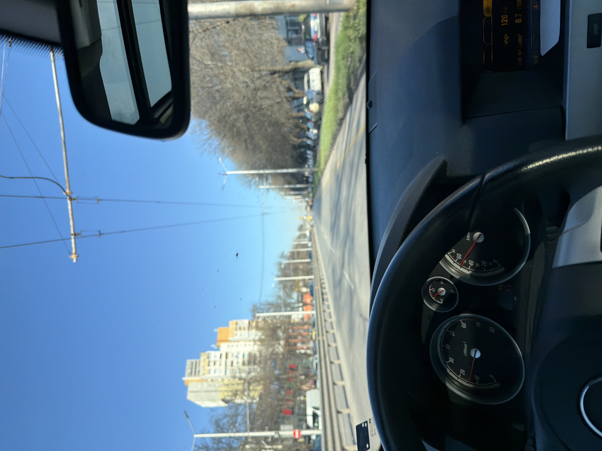 The image depicts a view from the interior of a car driving on a city street in Sofia, Bulgaria. The dashboard and steering wheel are visible in the foreground, obscuring the view of the road ahead. The speedometer and tachometer are visible, but unreadable in terms of current speed or RPM.
Through the windshield, a straight road lined with trees and buildings is seen, including a tall apartment building with yellow balconies on the left. Overhead trolley wires are strung. Other vehicles are parked along the right side of the road. A street display shows that the car's range is 126 km, and the consumption is 6.7 l/100 km.