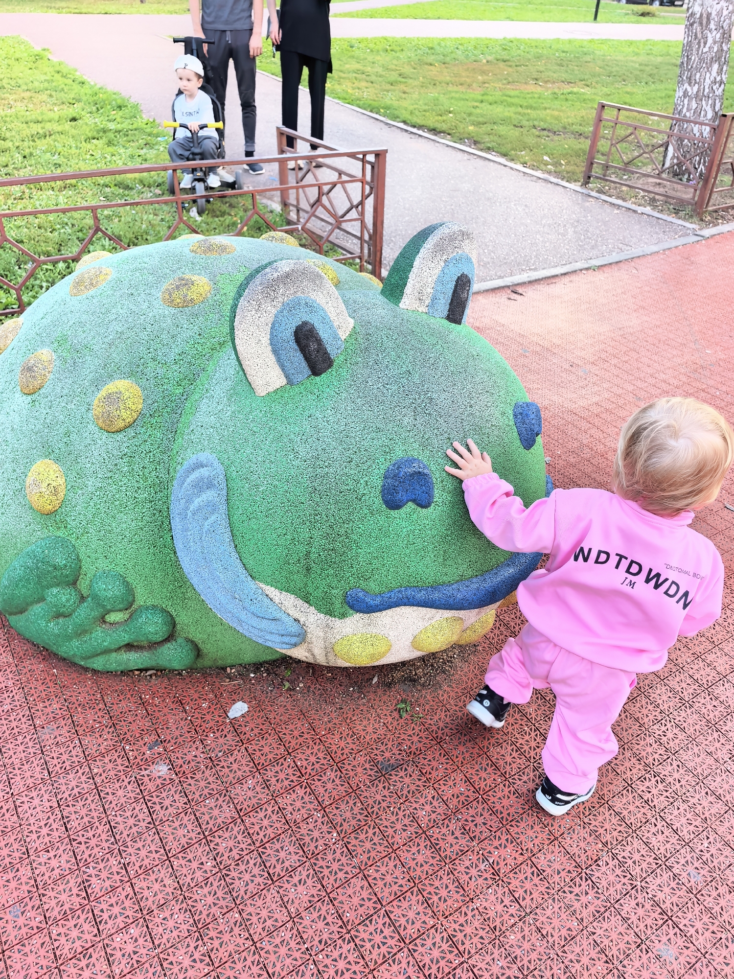 In Sterlitamak, Russia, a young child in a pink tracksuit touches a large, green, frog-shaped playground structure. The frog sculpture is textured and has yellow, blue, and gray accents. The ground is a red, rubberized surface with a patterned texture. In the background, a toddler sits on a tricycle, and two adults stand behind a fence. The environment appears to be a park or playground with green grass and a paved walkway.