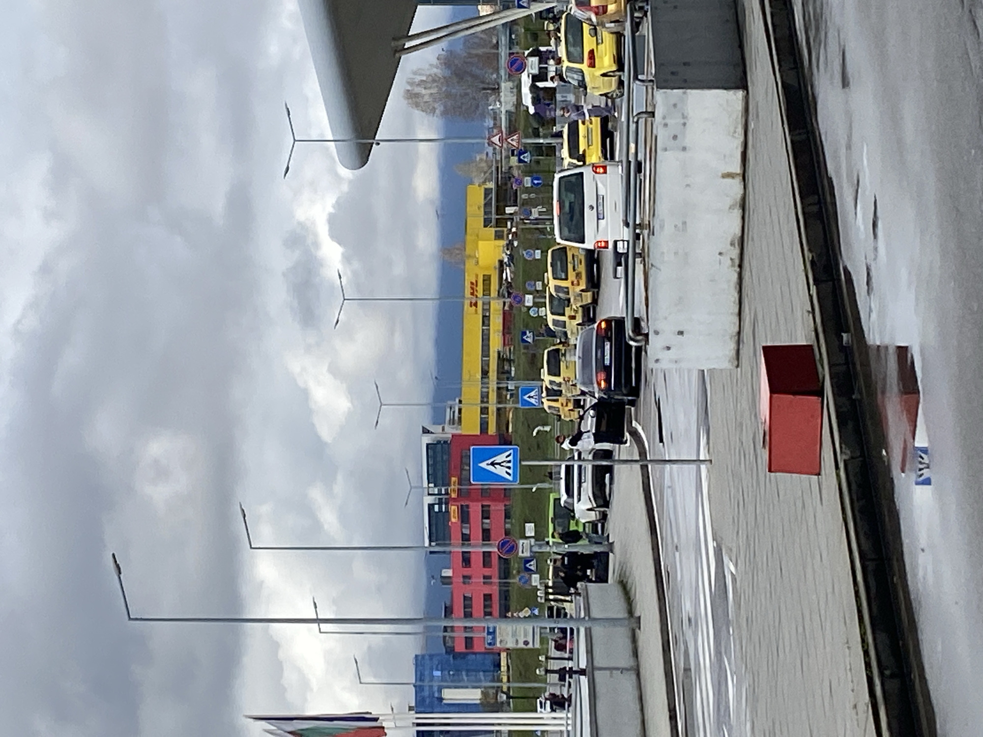 The image shows a street scene near Sofia Airport in Bulgaria. A line of yellow taxis and other vehicles is visible, along with street signs and colorful buildings (blue, red, yellow). An overpass or part of the airport structure is in the top of the frame, and Bulgarian flags are in the lower left. The sky is overcast.