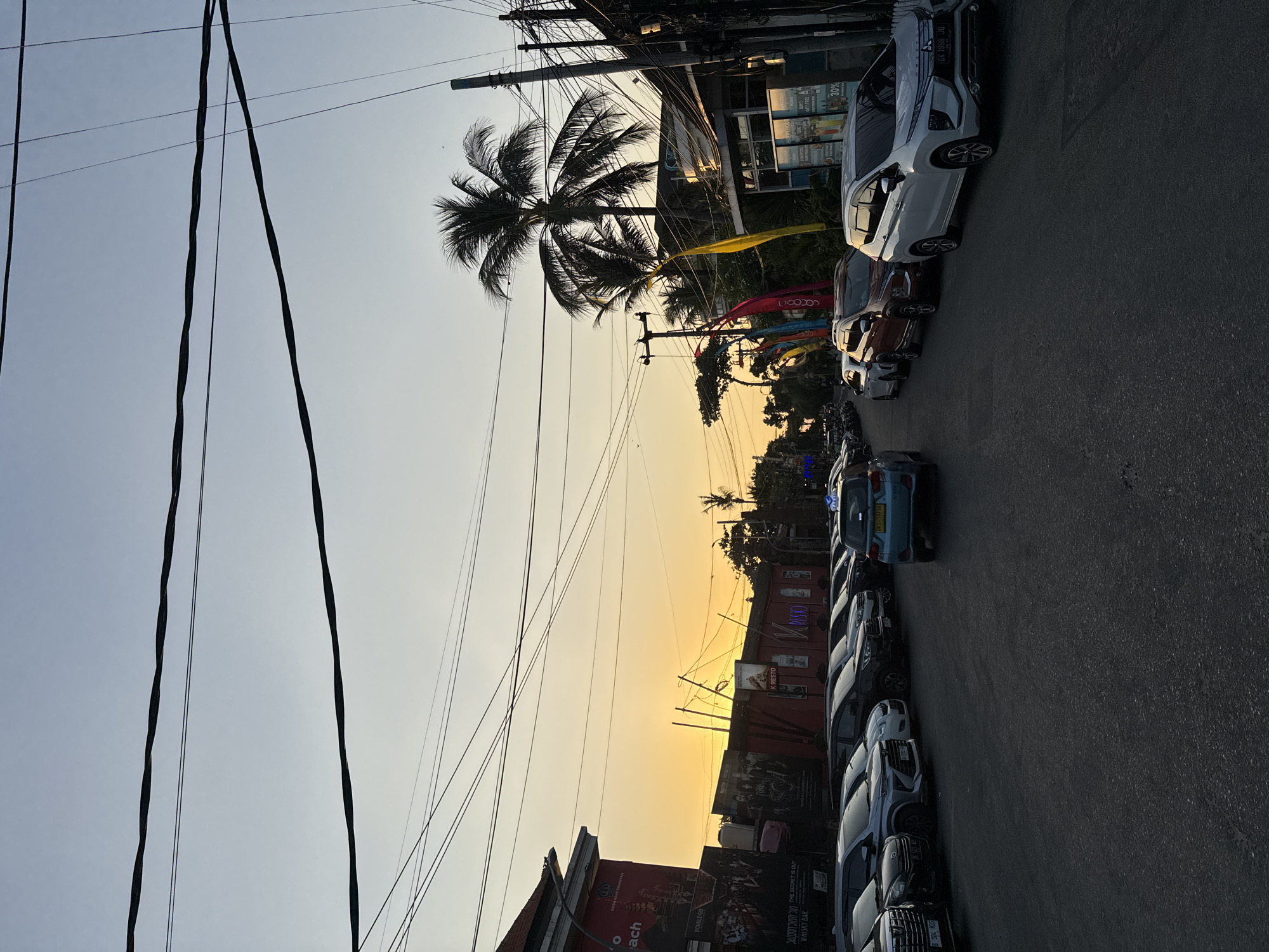 The image depicts a street scene in Kuta, Indonesia, during twilight. A row of cars is parked along the right side of the asphalt road, which is lined with buildings and palm trees. The sky is a soft gradient of pale blue and orange, indicating the setting sun. Overhead, a complex network of electrical wires crisscrosses the frame.
On the left side of the street, a building displays large posters with text that is partially legible, including "DESA ADA SEMARAK," "BEACH," and "WHISKY BAR." Further down the street, several shops and restaurants are visible, adorned with colorful flags and signage, such as "K RESTO."
The cars in the foreground and middle ground are mostly SUVs and sedans, parked parallel to the road. A single light blue car is in motion in the center of the road, heading away from the viewer. The overall atmosphere is that of a bustling commercial area as evening approaches.