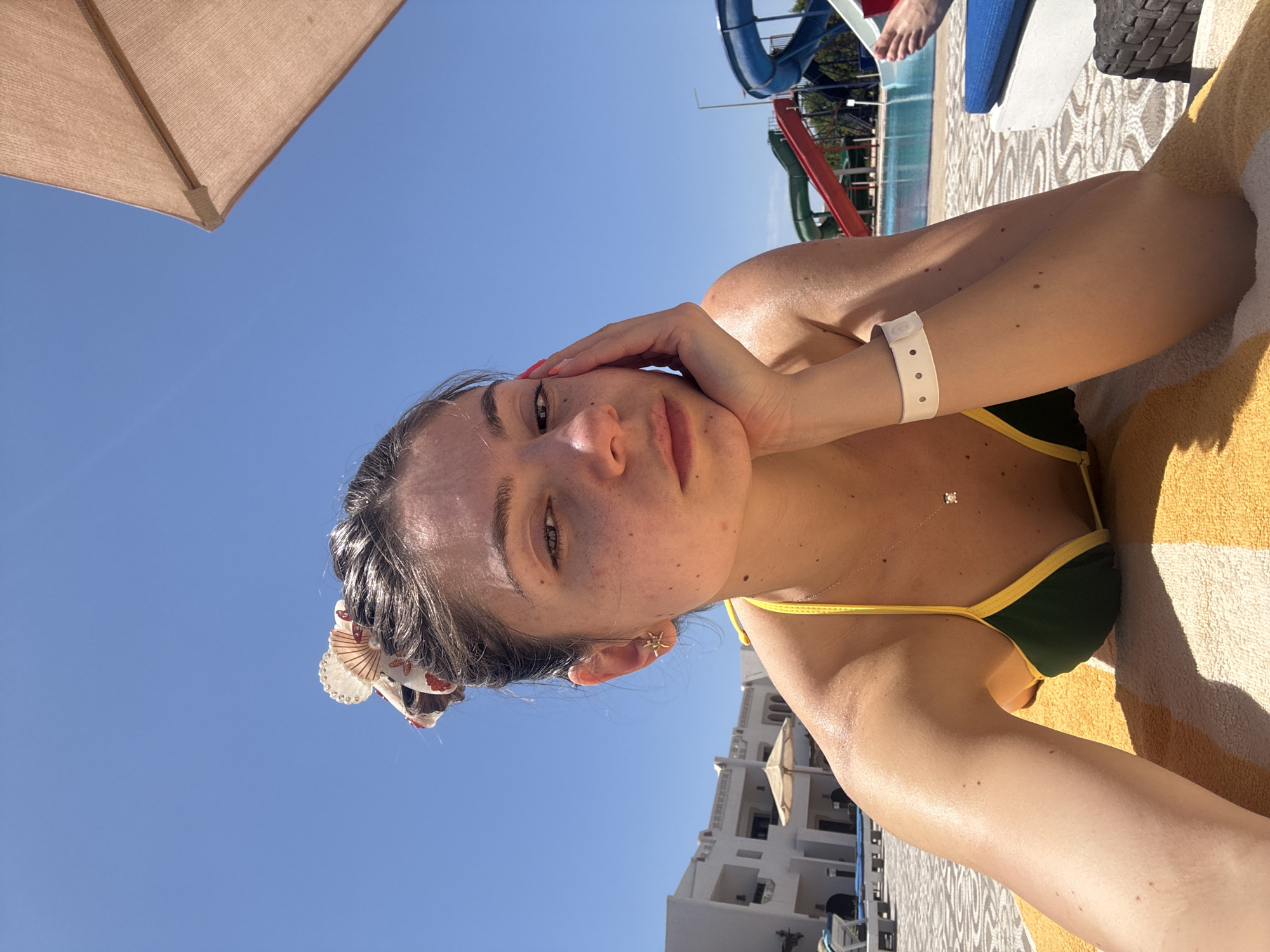 A portrait-oriented, eye-level photograph depicts a fair-skinned woman lying on a yellow and white striped towel under a brown parasol. Her dark hair is slicked back, adorned with a decorative shell hair clip. She is wearing a dark green bikini with yellow trim. Her left hand cradles her face, revealing an earring and a white wristband with holes. A delicate necklace with a small pendant is visible on her chest. In the background, a water park structure with blue and green slides is partially visible, along with a white building and a clear blue sky. The location appears to be a resort, but the specific city is not discernible from the visual information.