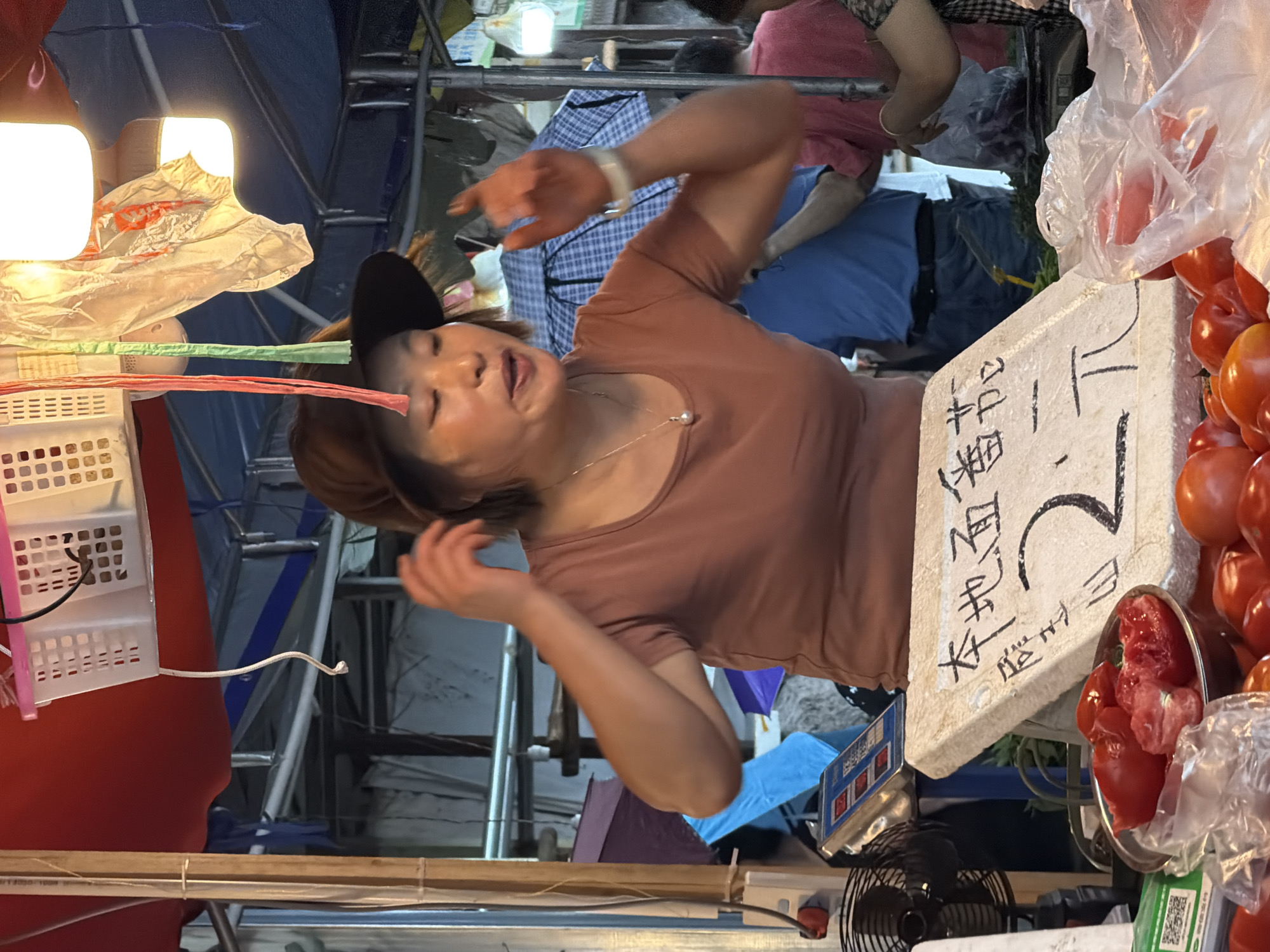 A market scene in Jiangjin, China, centers on a female vendor behind a stall laden with tomatoes. The woman wears a brown short-sleeved top and a dark brown wide-brimmed hat. A silver chain necklace with a single pearl pendant adorns her neck, and a white wristband is visible on her right wrist. Her mouth is open, and her hands are raised on either side of her head, indicative of speech or a gesture.
On the counter in front of her, a white styrofoam sign displays handwritten Chinese characters "本地面番茄" (Local Flat Tomatoes) and a price of "2元" (2 Yuan). To the left of the sign, a metal bowl contains several red tomatoes, some appearing cut or damaged. To the right and in the foreground, a pile of whole red tomatoes is visible, with some enclosed in clear plastic bags. An electric fan and a digital scale with a blue display are situated behind the tomato display on the left, alongside a green QR code sticker.
The market environment features an overhead red canopy and structural elements including metal poles and white plastic crates hanging from above. Illumination is provided by overhead lights, some appearing bare or covered with plastic bags. Colorful fabric strips also hang from the ceiling. In the background, other individuals are visible, including a man in a blue t-shirt and dark pants, and a person in a red shirt partially obscured while interacting with goods. Several umbrellas, including a blue and white checkered one, are scattered throughout the background. The scene depicts a lively and functional commercial setting.
