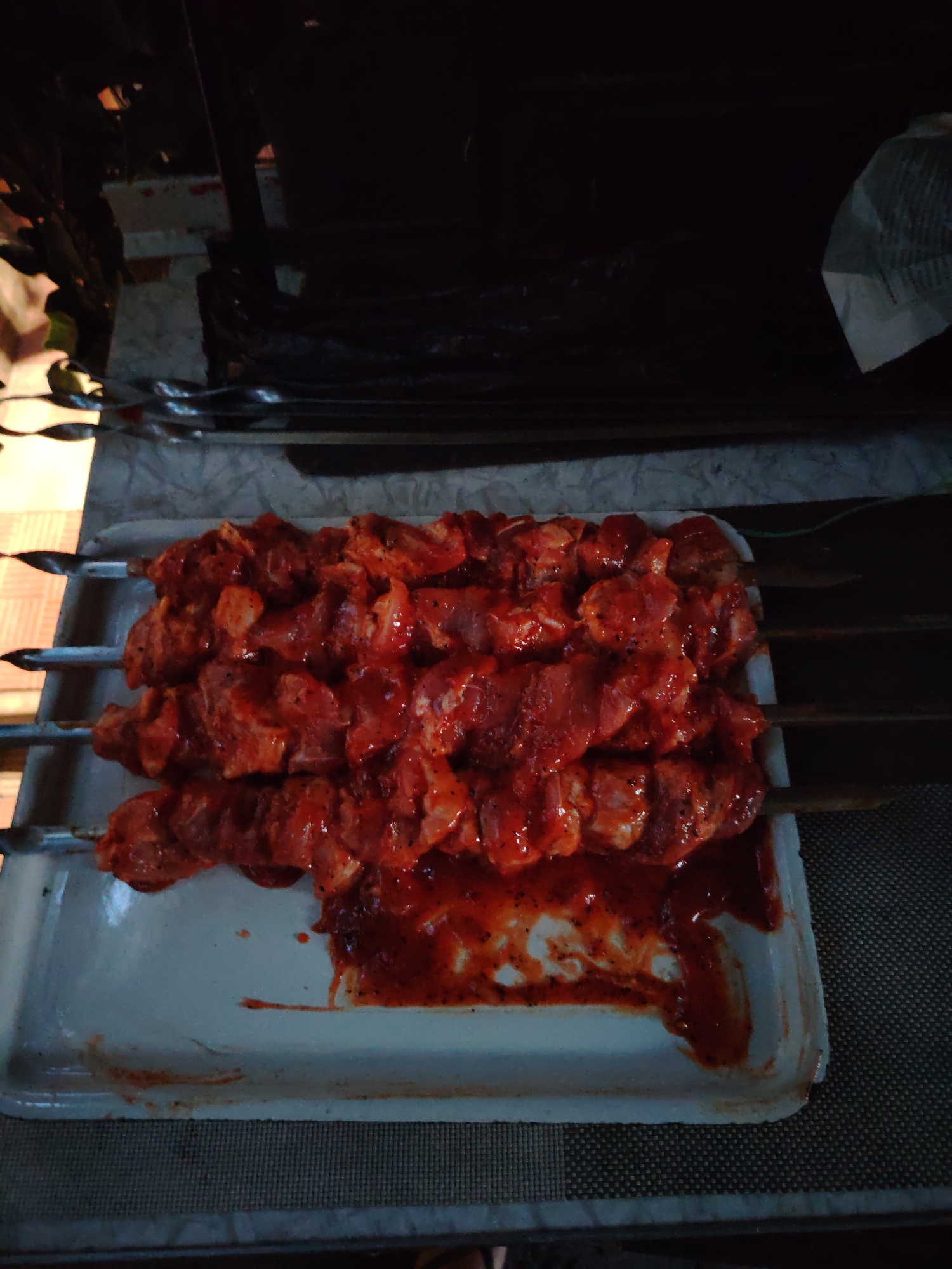 A tray of skewered raw meat, coated in a red marinade, sits on a patterned surface. The meat is arranged in four rows on metal skewers, with some marinade pooled in the tray. The background is dark and out of focus, showing hints of plants and a crumpled white object. The image appears to have been taken indoors.