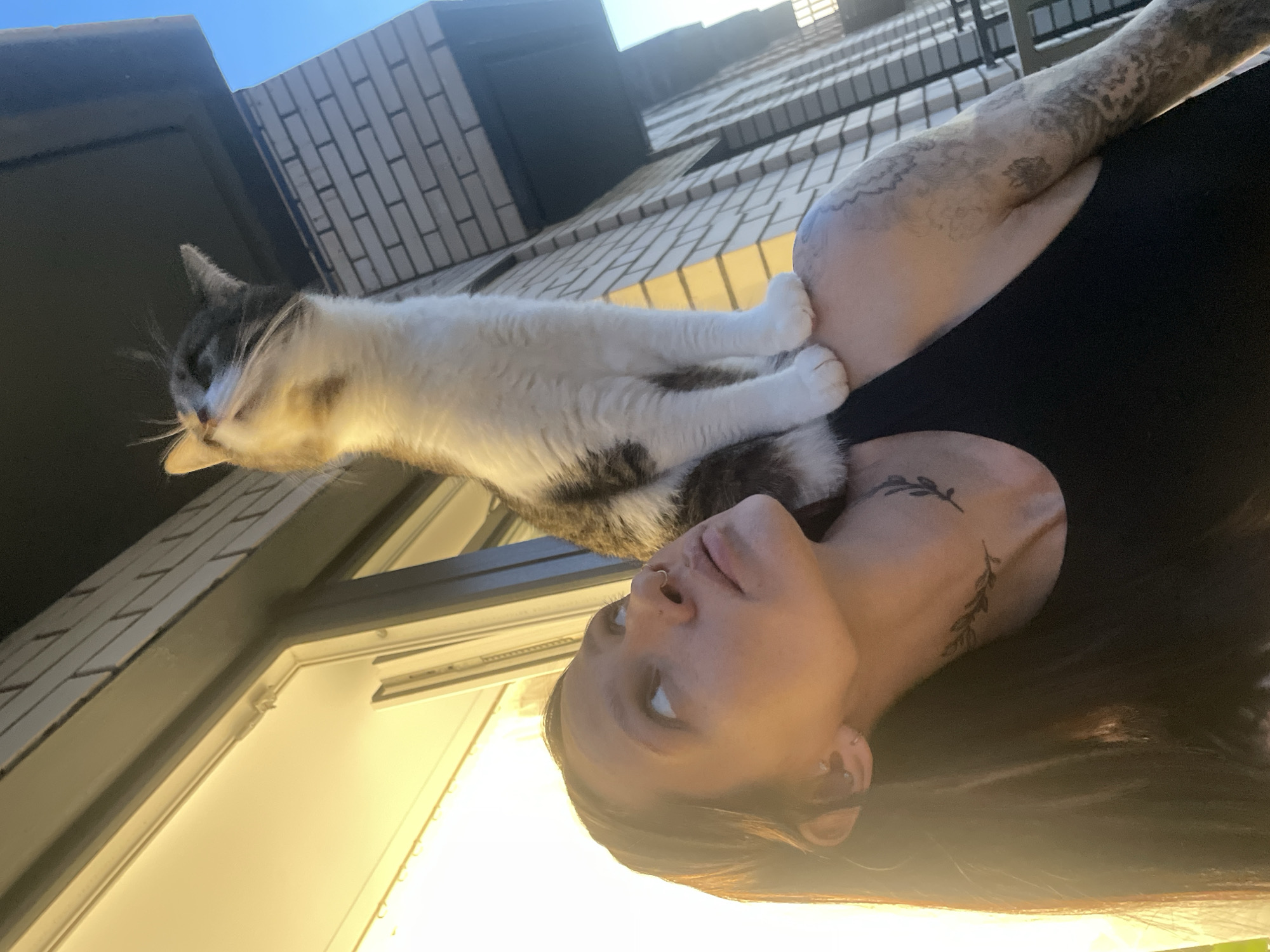 A woman stands outdoors in Pervomaysky, Russia, with a cat draped around her shoulders. The woman is facing upwards, her head tilted back slightly. She has fair skin, dark hair, and tattoos on her neck and arm. A nose ring is visible. The cat is white with gray and brown markings. The background features a brick building and a clear blue sky. Sunlight is diffused across the lower portion of the image.