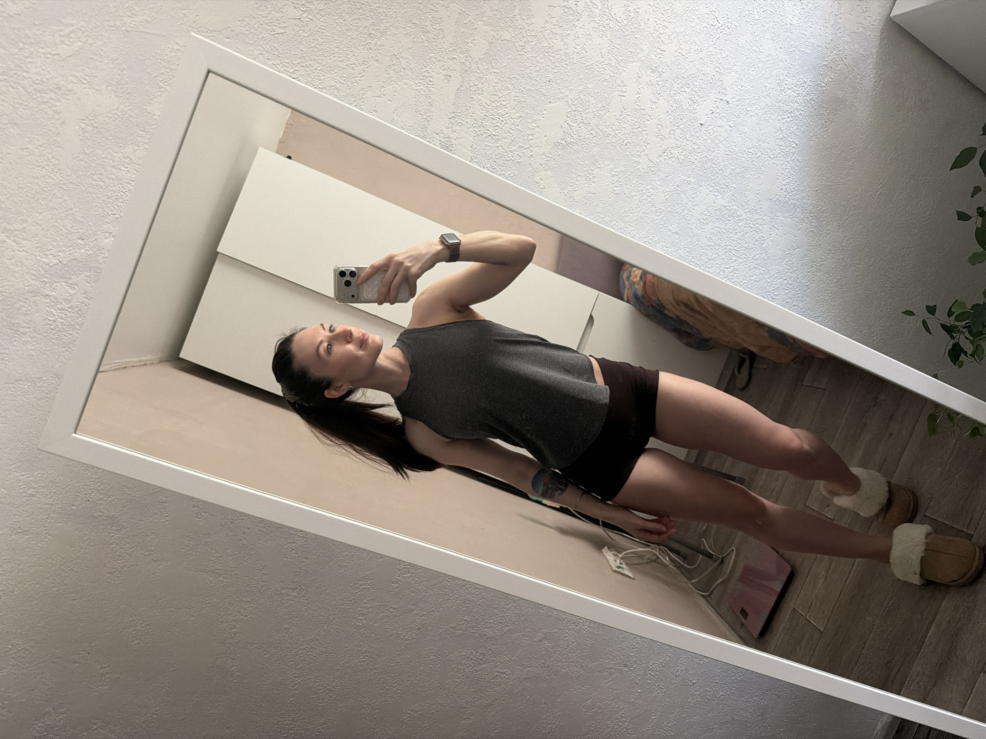 A young woman, likely in her 20s or 30s, stands in an indoor setting in Perm, Russia, taking a full-body mirror selfie. She is dressed casually in a dark grey sleeveless top, dark shorts, and light brown Ugg-style slippers with white fur lining. Her dark hair is pulled back in a ponytail, and she wears a smartwatch on her left wrist. She holds a smartphone, possibly an iPhone, in her right hand, positioned to capture her reflection. A colorful tattoo is visible on her upper right arm.
The mirror, framed in white, is leaning against a textured white wall. The reflection reveals a tidy room with white cabinet-like furniture, a pink weight scale on the floor near her feet, and white charging cables. A green leafy plant is partially visible in the top right corner of the mirror's reflection. The floor appears to be light-colored wood or laminate. The woman maintains a neutral to slightly smiling expression, looking towards the phone's screen. The lighting suggests it is daytime indoors. No discernible text is visible on her clothing or the objects, though the phone case has a patterned texture.