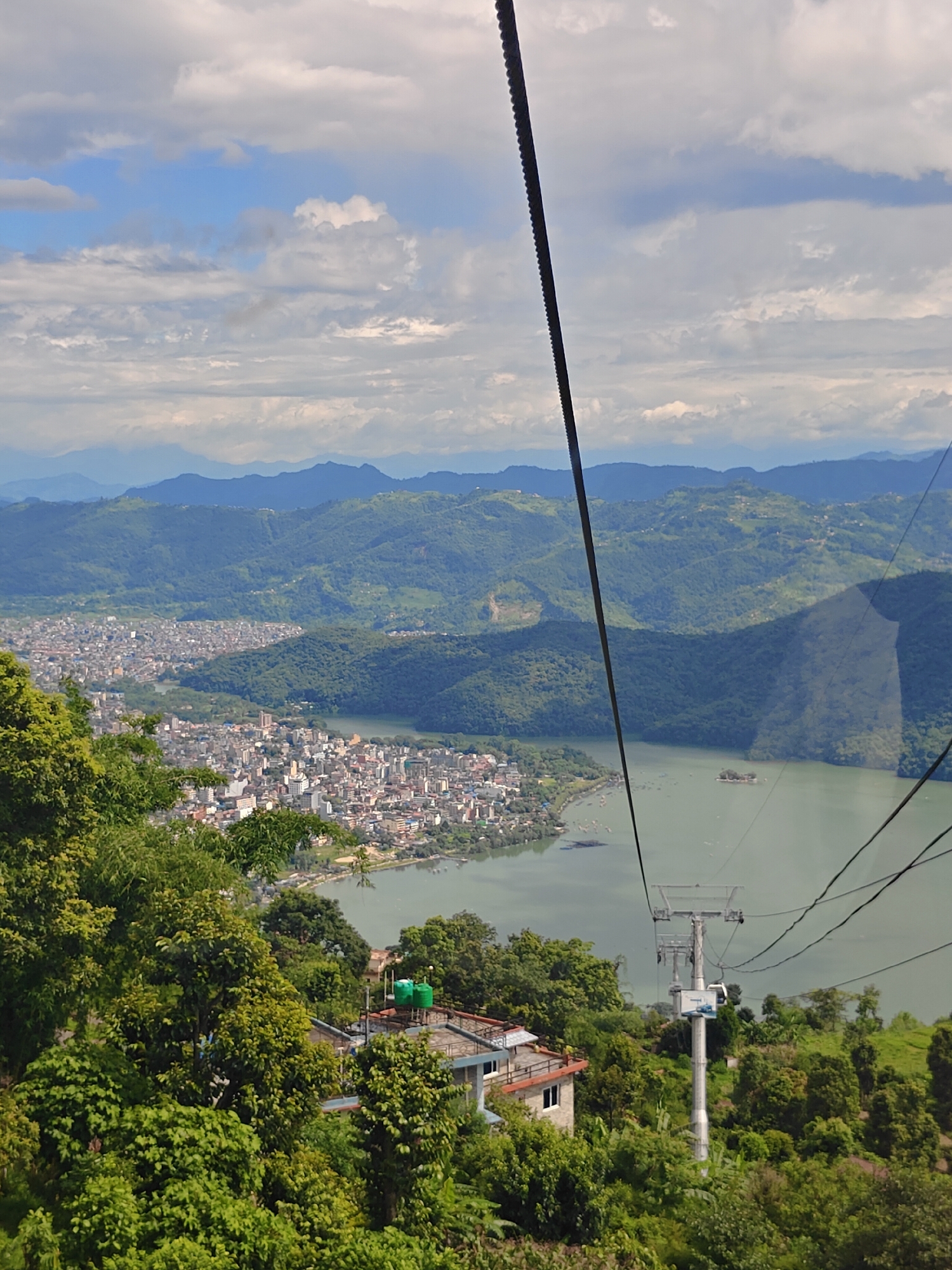 This is a vertical, eye-level shot taken from a cable car ascending a mountain overlooking the city of Pokhara, Nepal. The foreground is dominated by lush green trees, with a portion of a building structure featuring water tanks visible on the right. A cable car support tower is also prominent in the mid-ground, with cable car lines extending upwards and diagonally across the frame.
The mid-ground of the image reveals a sprawling cityscape of Pokhara situated along the edge of a large, calm lake, identified as Phewa Lake. The city is characterized by numerous buildings of varying heights, clustered together. The lake itself is a tranquil, light green body of water, with several small boats scattered across its surface. A small island with a temple is visible in the center of the lake.
The background showcases a series of rolling, verdant hills and mountains, layered in shades of green and blue due to atmospheric perspective. The distant mountains appear as soft, hazy silhouettes against a sky filled with scattered white clouds. The cable car itself is a dominant element in the composition, its thick cable running diagonally from the top left corner towards the center.