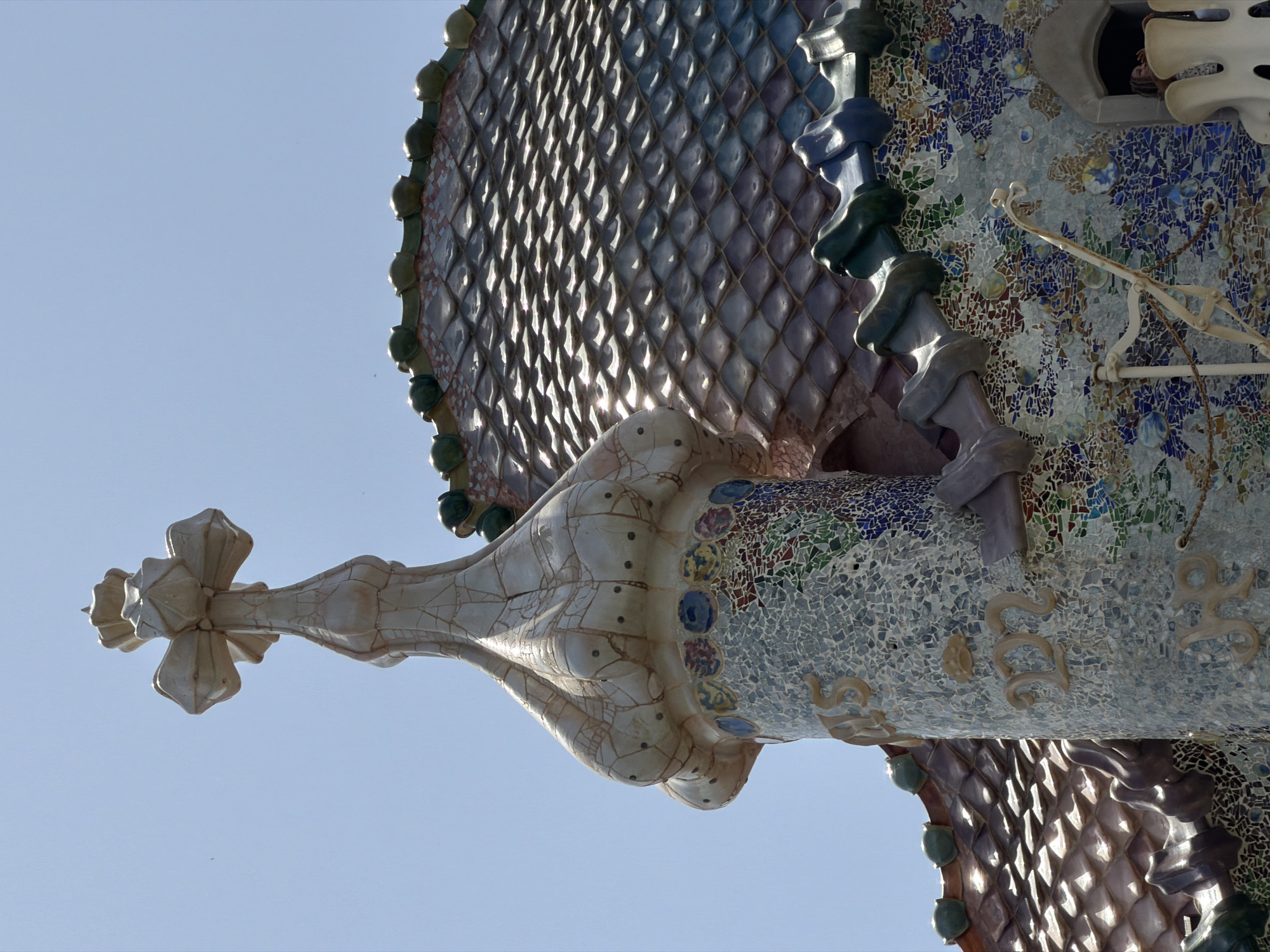 This is a close-up, eye-level shot of the roof and chimney of Casa Batlló, a building located on Passeig de Gràcia in the Eixample district of Barcelona, Spain, designed by architect Antoni Gaudí. The image focuses on the top section of a chimney tower, which is covered in intricate multicolored mosaic tiles. The tower is topped with a four-armed cross, also tiled in a light, textured material that appears to be cracking in places. To the right of the chimney, a portion of the building's distinctive roof, resembling dragon scales, is visible. The roof tiles are a mosaic of earth tones and blues, with rounded, bulbous green elements forming the ridge. The sky is clear and a pale blue. The mosaic on the tower features patterns of blue, green, red, and yellow fragments, interspersed with larger, round, disc-like elements in various colors. Inscriptions in a golden, raised font are visible on the mosaic-covered tower, including what appears to be "MAS" and "222". A decorative white metal structure with a chain is also partially visible in the lower right section of the image, near a window opening from which a person's head and shoulders can be seen. The overall impression is one of ornate, organic architectural design.