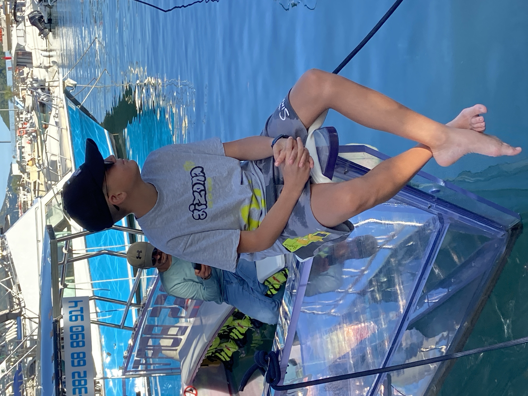 An overhead, slightly tilted shot captures a young person relaxing in a transparent boat or kayak on clear, calm water in Budva, Montenegro. The main subject, a boy, is lying on his back, looking upwards, with his hands clasped over his stomach. He wears a grey t-shirt with stylized black and yellow text (possibly "CROZ"), grey shorts, and a black baseball cap. His bare feet are visible, elevated above the transparent hull. A dark wristband or watch is on his left wrist.
The transparent vessel reveals the blue-green water beneath and around it, allowing light to refract and reflect, creating patterns on the boat's interior surfaces. Stacked yellow and black buoyancy aids or life vests are visible inside the boat, to the left of the main subject.
Partially visible in the background, an adult male wearing a light-colored long-sleeved shirt and a baseball cap with a prominent "H" logo is seated within the same transparent boat, holding a mobile phone.
The scene takes place on a bright, sunny day, suggested by the strong light and clear reflections on the water. In the background, a marina or harbor setting is evident, with several white boats docked along a quay. Distant land or mountains line the horizon. On the side of one boat or structure in the mid-ground, the alphanumeric text "HT6 068 89 28E" is clearly visible. The overall mood is one of relaxation and leisure on the water.