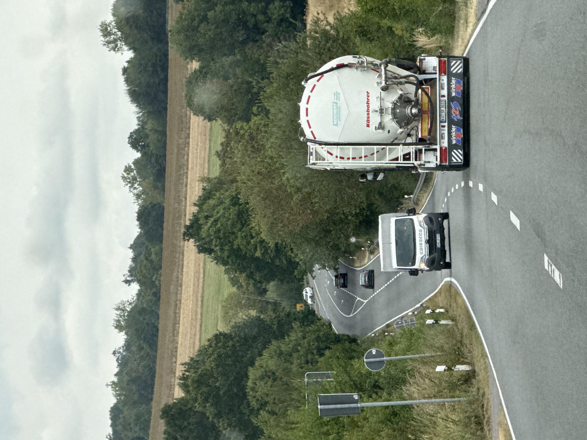 The image shows a winding road in a rural setting, likely in or around Bad Sachsa, Germany. The sky is overcast. The road curves to the left in the foreground, with a white van in the right lane. Further ahead, a large white tanker truck with "Kässbohrer" and "REICHERT" printed on it is in the right lane, followed by a black car. In the distance, another black car is visible, and beyond that, a white car. The road is bordered by trees and vegetation on both sides. Fields are visible in the background, suggesting a countryside location. Traffic signs and road markings are present.