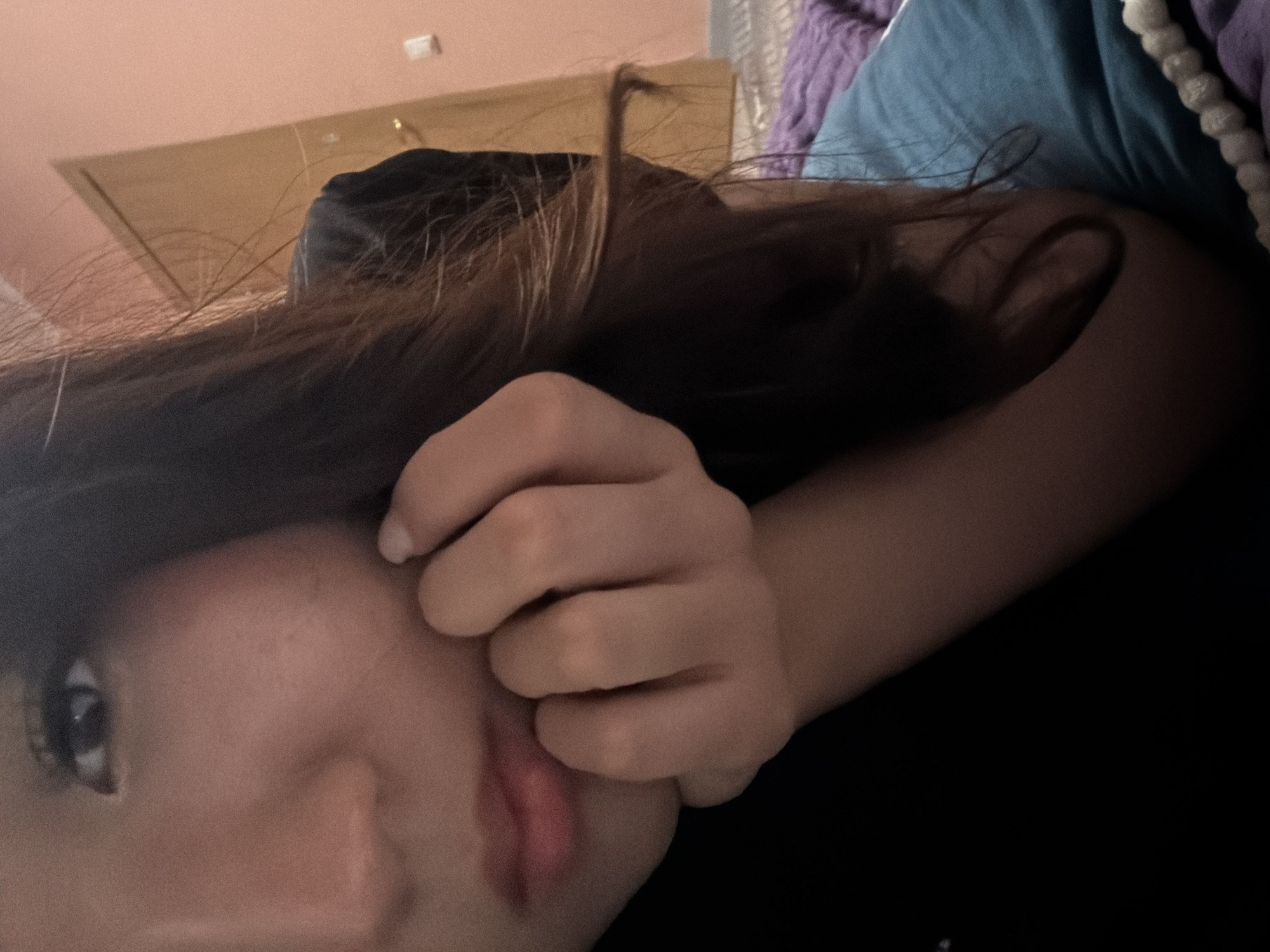 A close-up, low-angle shot shows a person's face partially obscured by their hand and dark brown hair. The person is lying down, resting their head on their hand. A pink wall and a light-colored door are visible in the background, suggesting an indoor setting, likely a bedroom. The lighting is dim, creating a soft, warm atmosphere. The person is wearing a black shirt. There are no visible signs of the city of Unknown, Russia, or any other specific location.