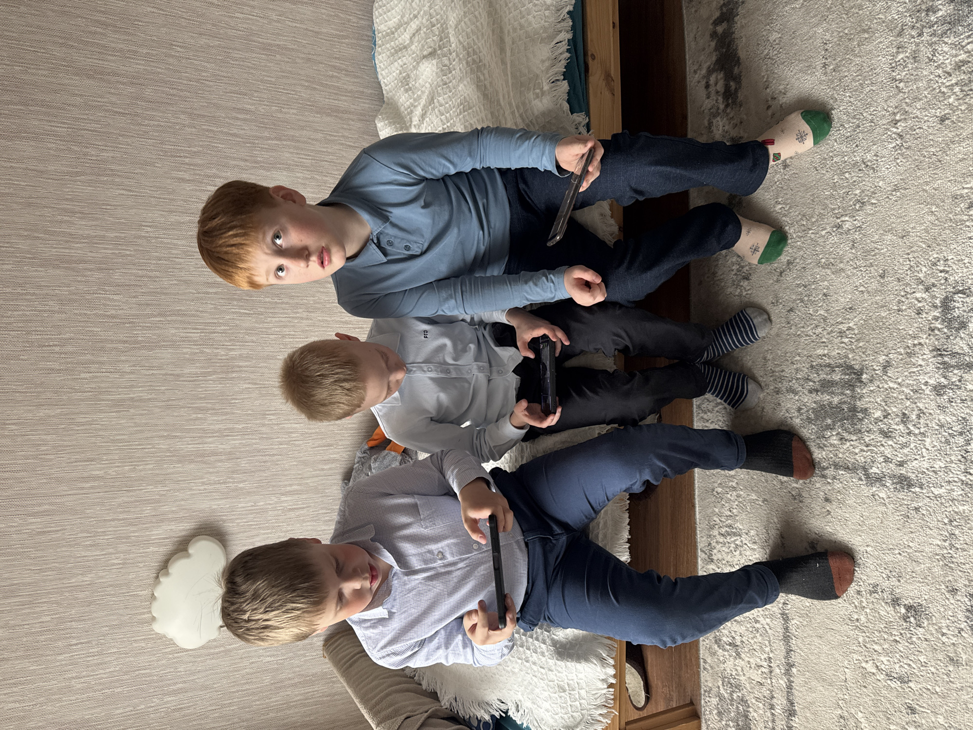 Three boys are sitting on a bed. The boy at the top has red hair and wears a blue long-sleeved shirt and dark pants. He is holding a tablet. He is wearing green and white patterned socks. The middle boy has blond hair, wears a white long-sleeved shirt and dark pants. He is holding a gaming controller. He is wearing blue and white striped socks. The boy at the bottom has blond hair and wears a white patterned button-down shirt and blue pants. He is holding a tablet. He is wearing black socks with brown toes. The bed has a wooden frame and a white textured bedspread. The carpet is light-colored with a mottled pattern. A white cloud-shaped object is on the floor to the left of the bed. The location is unspecified, and there is no indication that this image was taken in Votkinsk, Russia.
